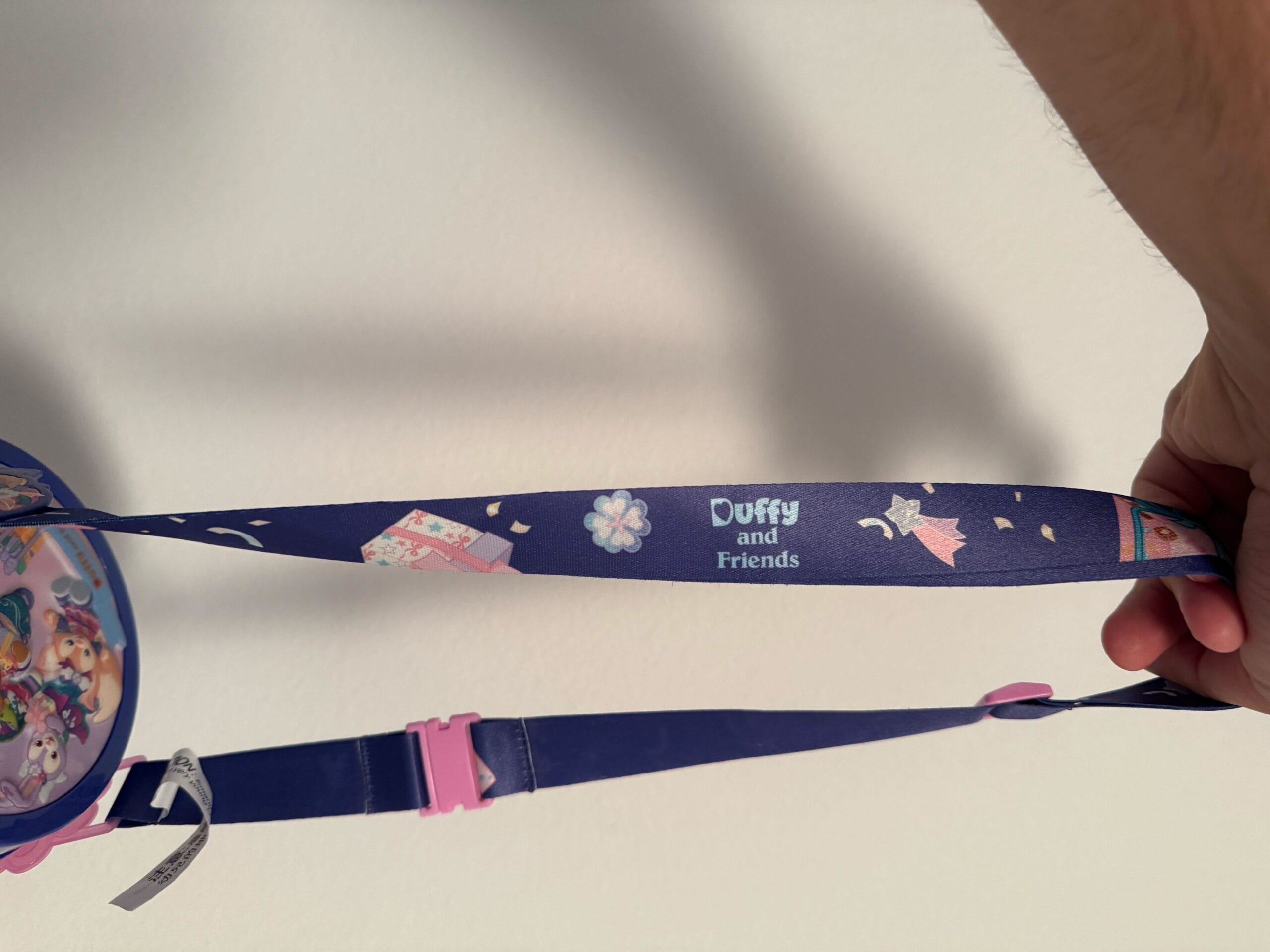 A hand holds a purple "Duffy and Friends" lanyard with cute designs and a pink buckle against a white wall.