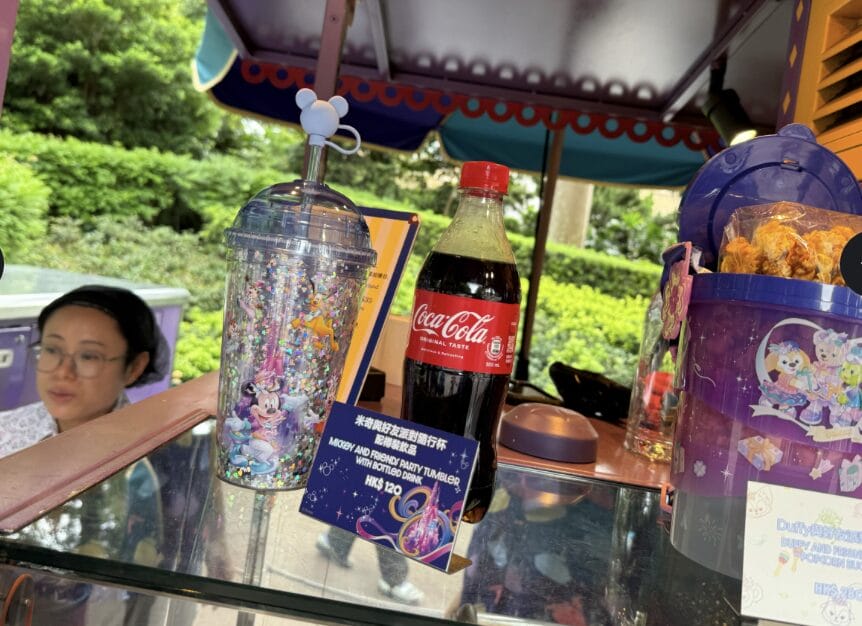 A Coca-Cola bottle, a Minnie Mouse cup, and snacks are displayed at an outdoor food cart, with a woman in the background.