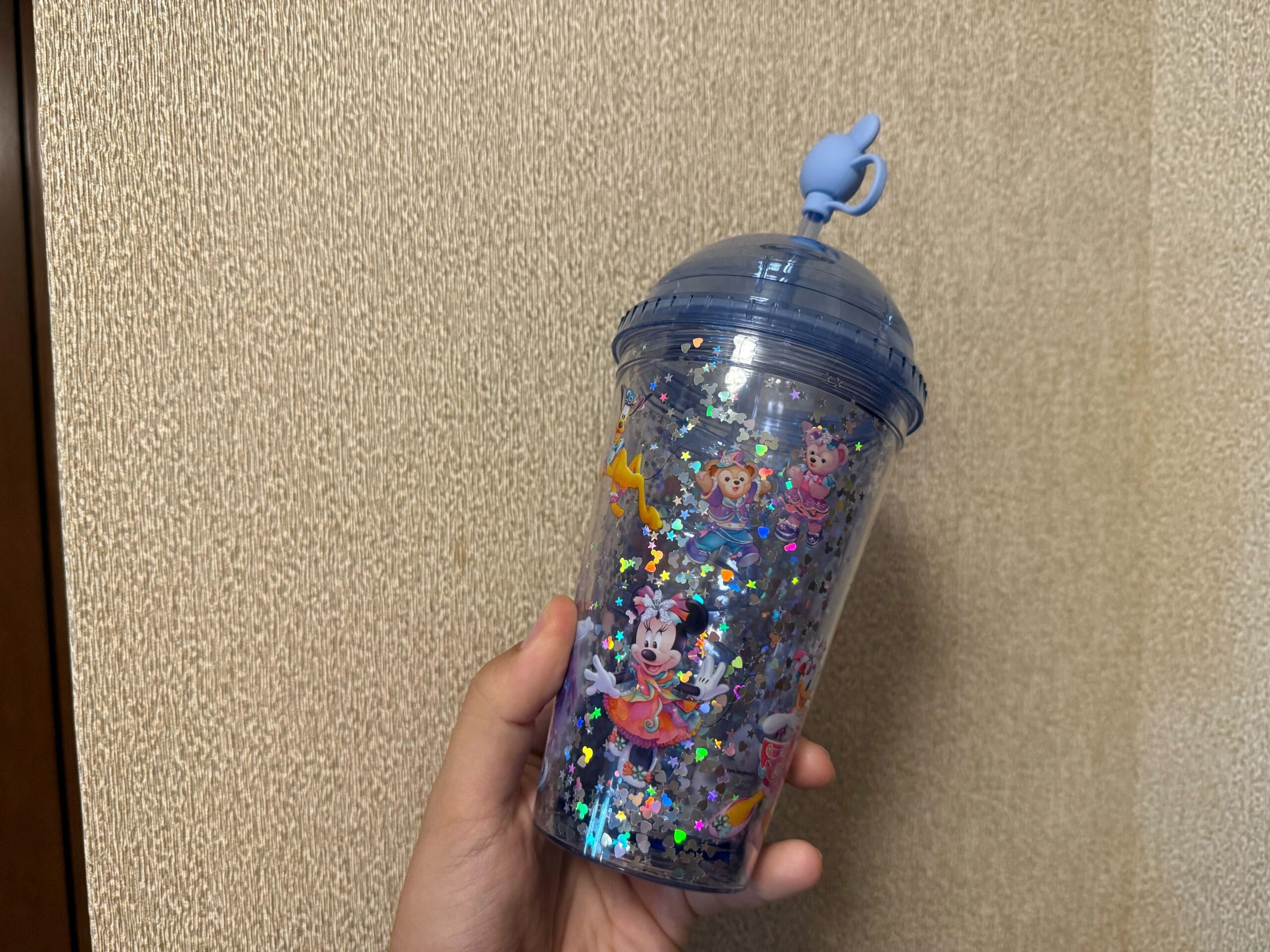 A hand holds a glittery cup with Disney characters and a blue Genie lamp-shaped lid against a beige wall.