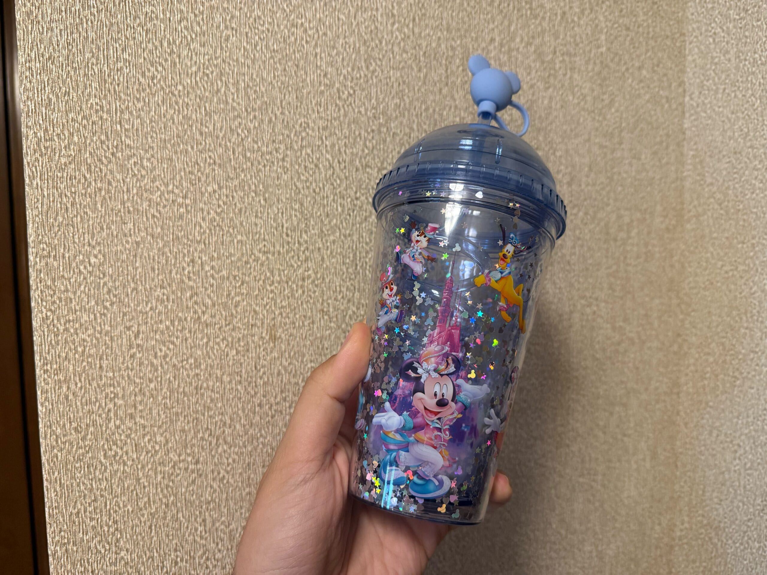 Hand holding a clear cup with Disney characters, blue lid, mouse-ear topper, and shiny star-shaped confetti inside.