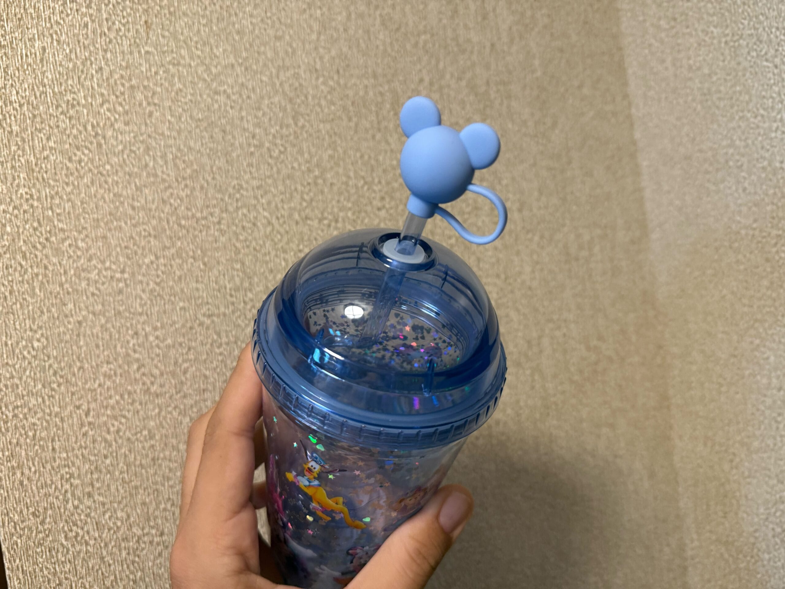 A hand holds a blue tumbler with a dome lid and a straw topped with a blue mouse-shaped figure.