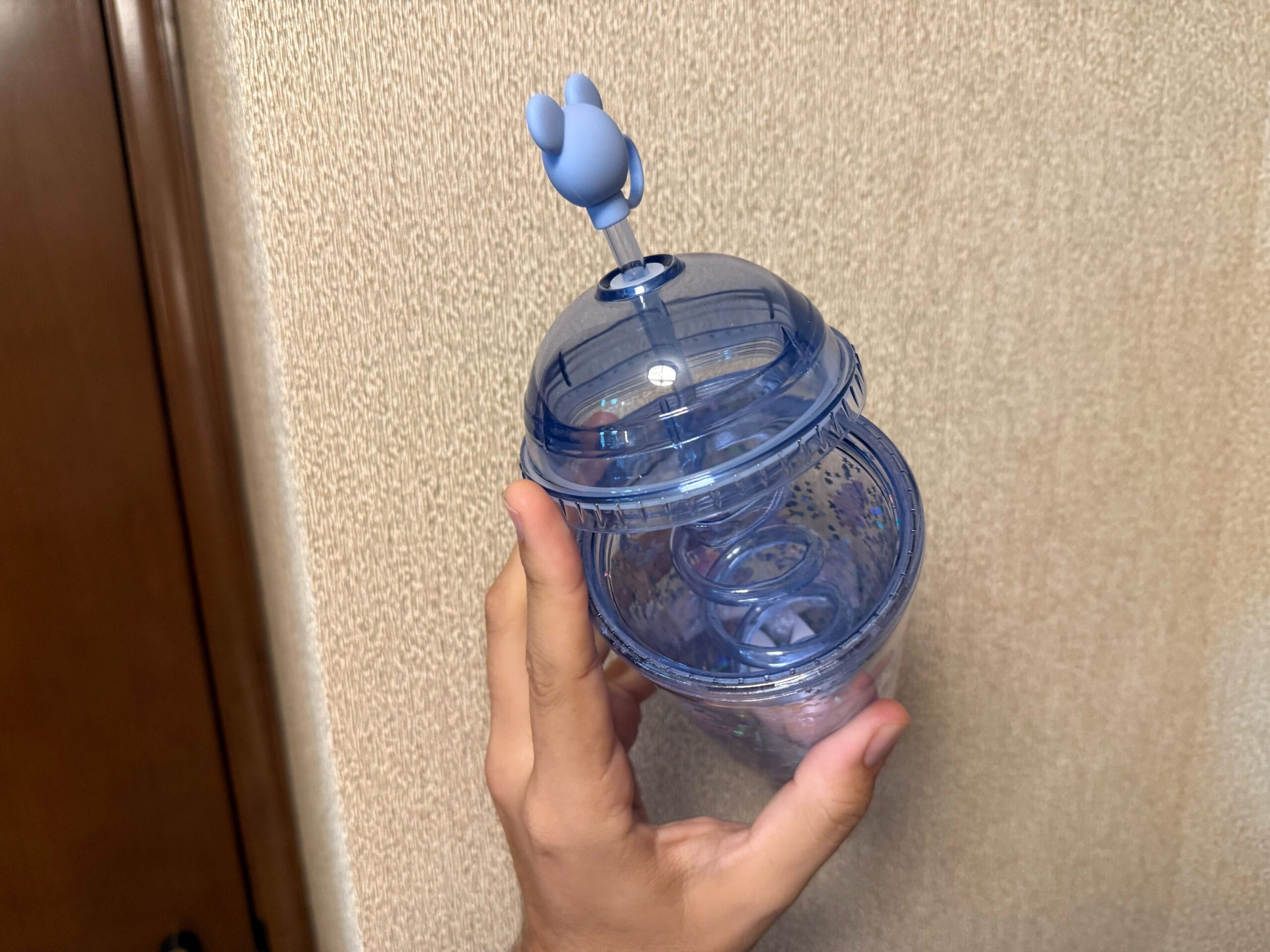 A hand holding a blue spiral tumbler cup with a bunny-shaped figure on the straw, against a beige wall.