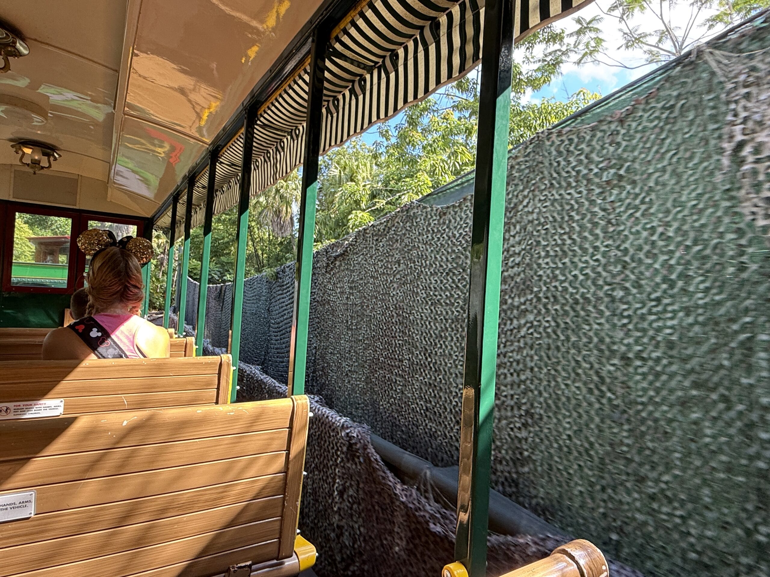 A person in Mickey Mouse ears rides a tram near Frontierland Station, passing green walls beneath a striped canopy.