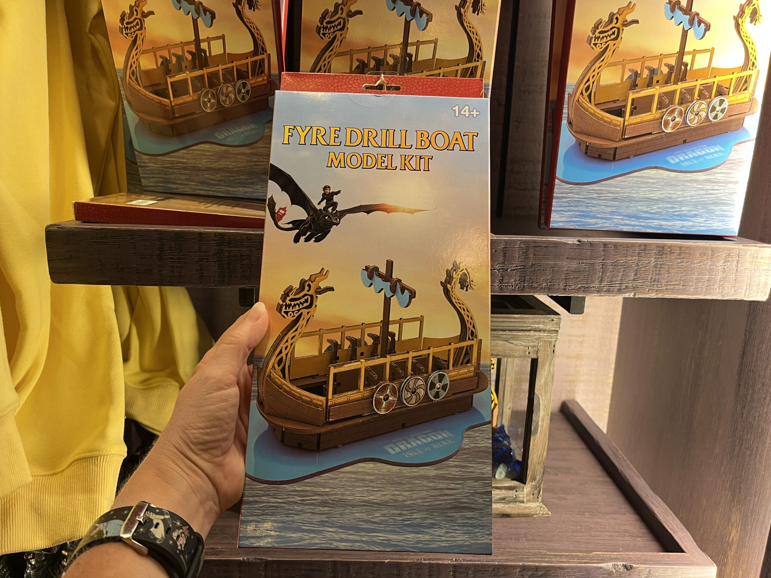 A hand holds a "Fyre Drill Boat Model Kit" from Hiccup's Work Shop, featuring a dragon boat image, in a store display.