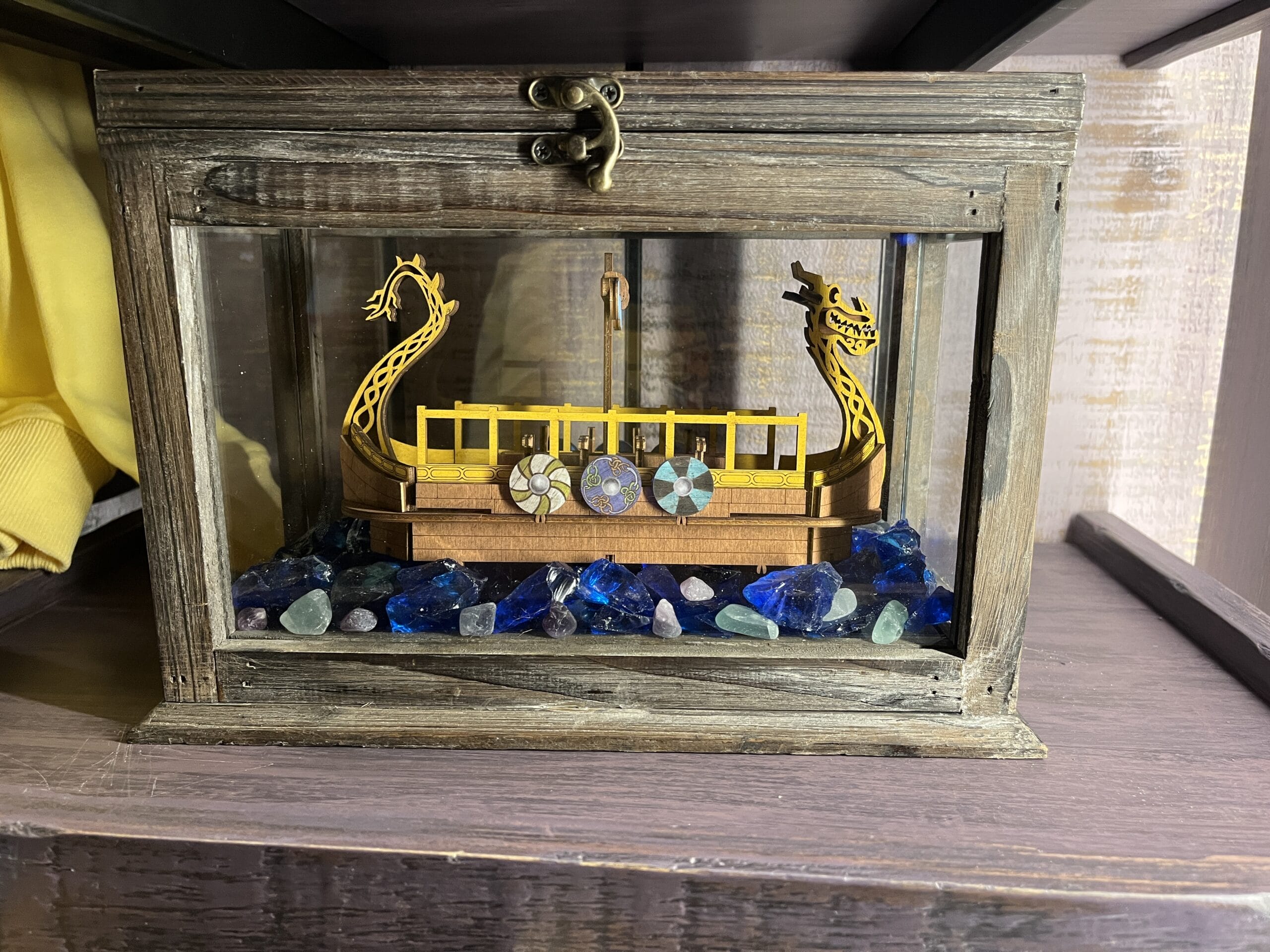 A wooden model Viking ship from Hiccup's Work Shop, with shields, in a glass case amid blue and green stones.