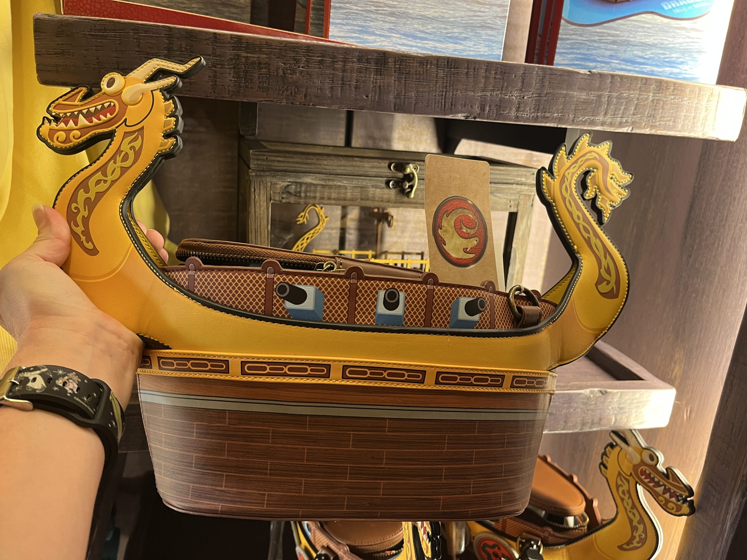 A hand holds a Viking ship-shaped purse from Hiccup's Work Shop, with yellow dragon heads and decorative details.