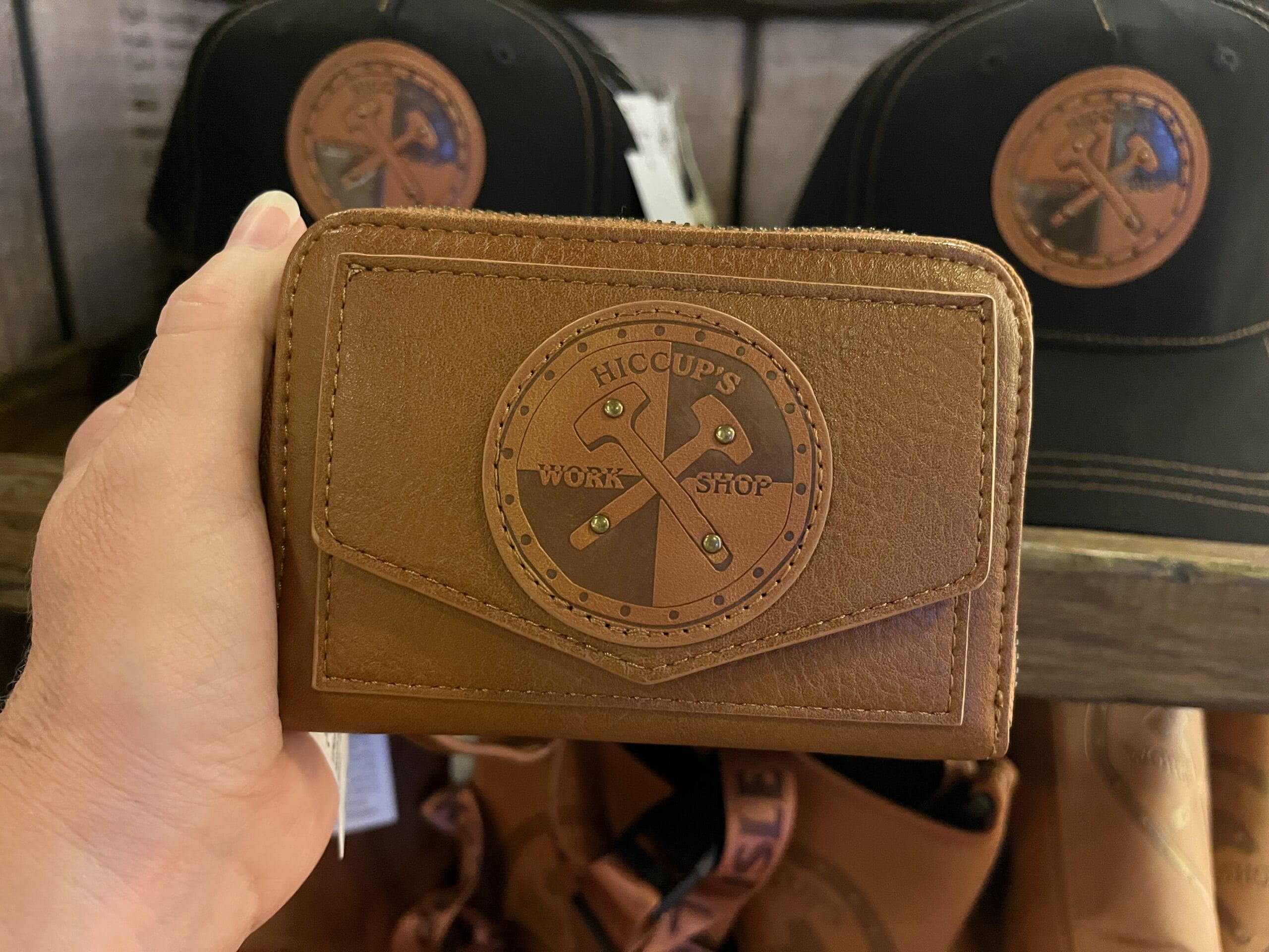 A hand holds a brown wallet labeled "Hiccup's Work Shop" with hats and bags in the background.