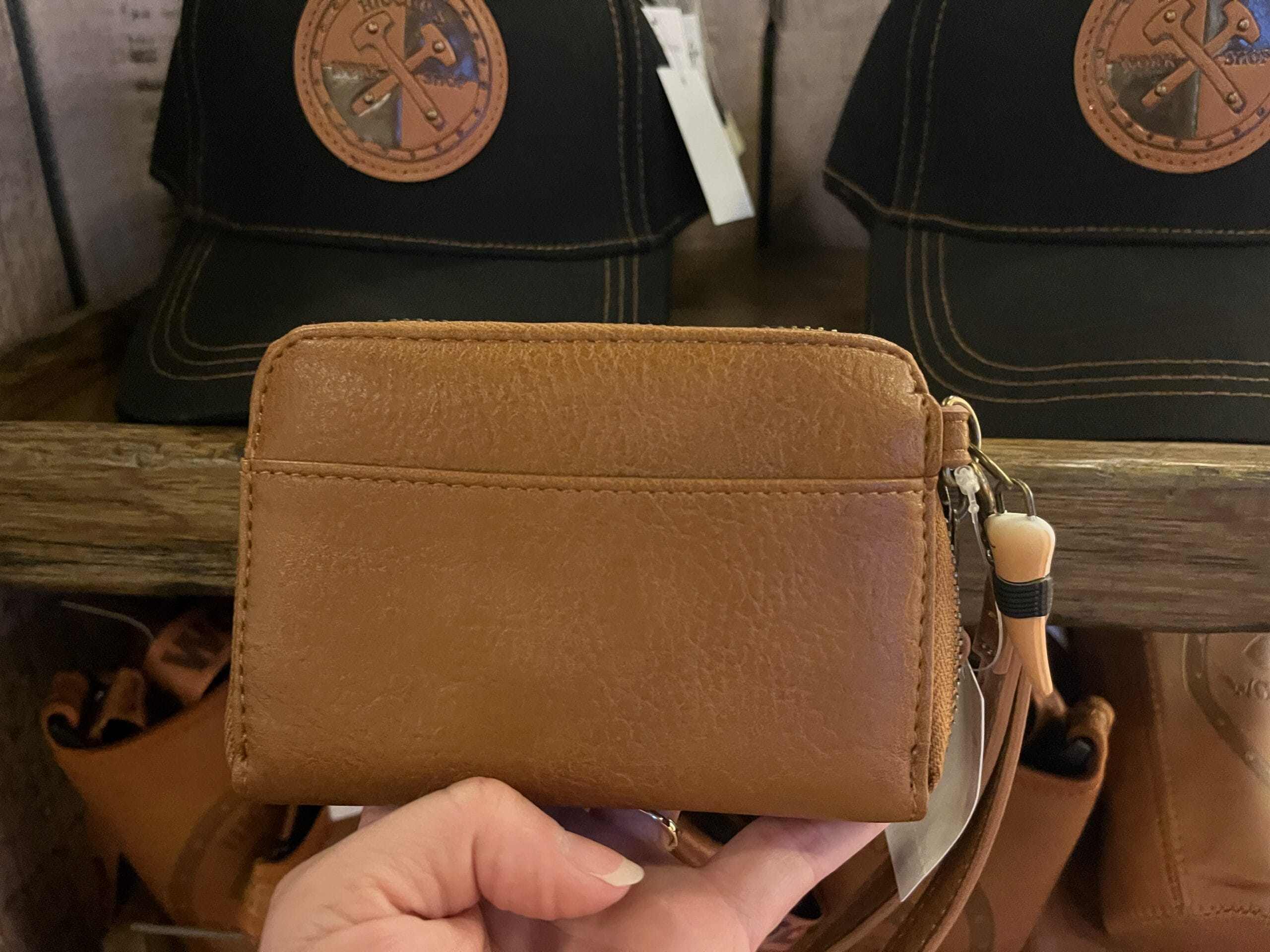 A hand holds a brown leather wallet in front of Hiccup's Work Shop hats with brown patches on a wooden shelf.