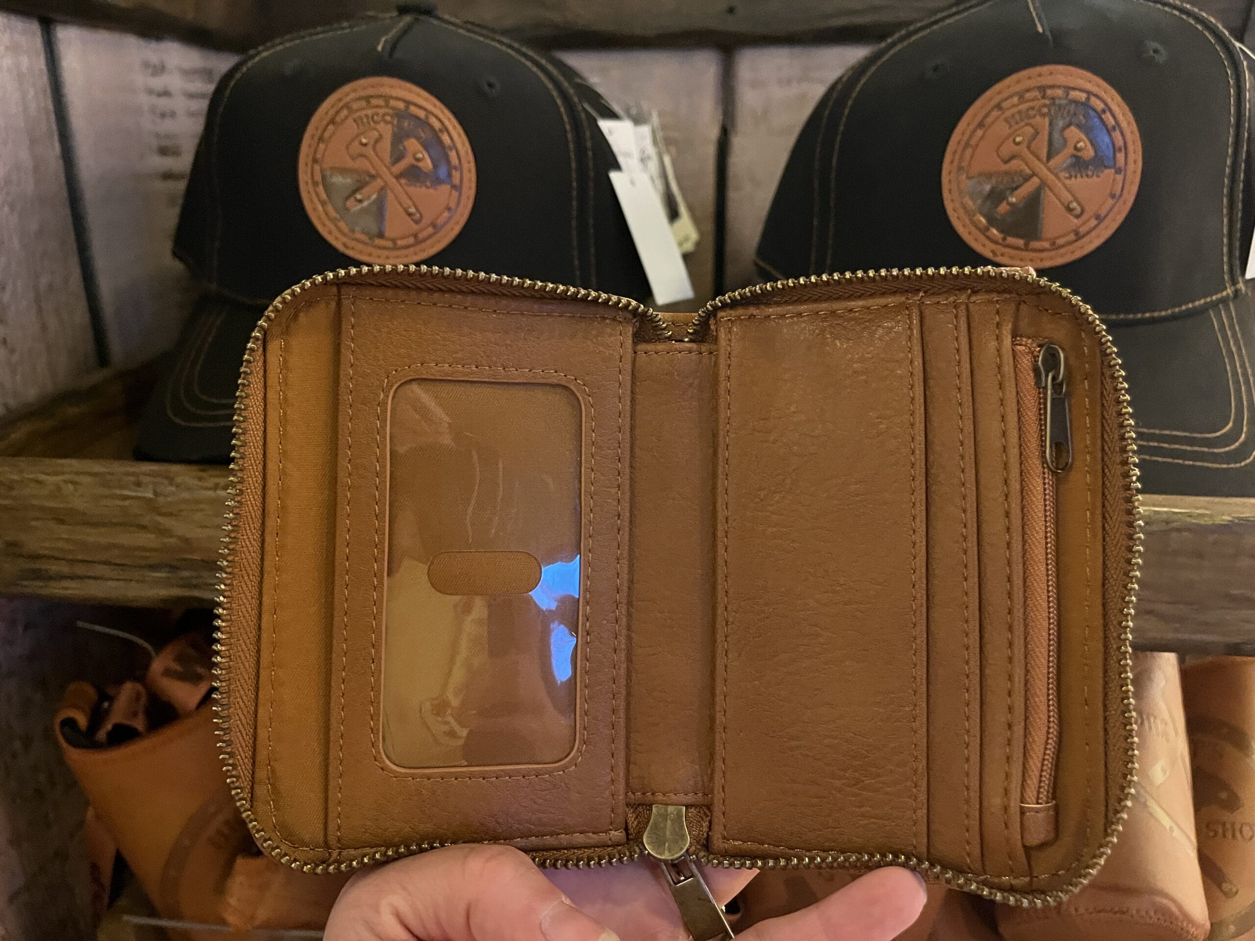 A hand holds open a brown leather wallet by Hiccup's Work Shop in front of black hats with leather patches on a wooden shelf.