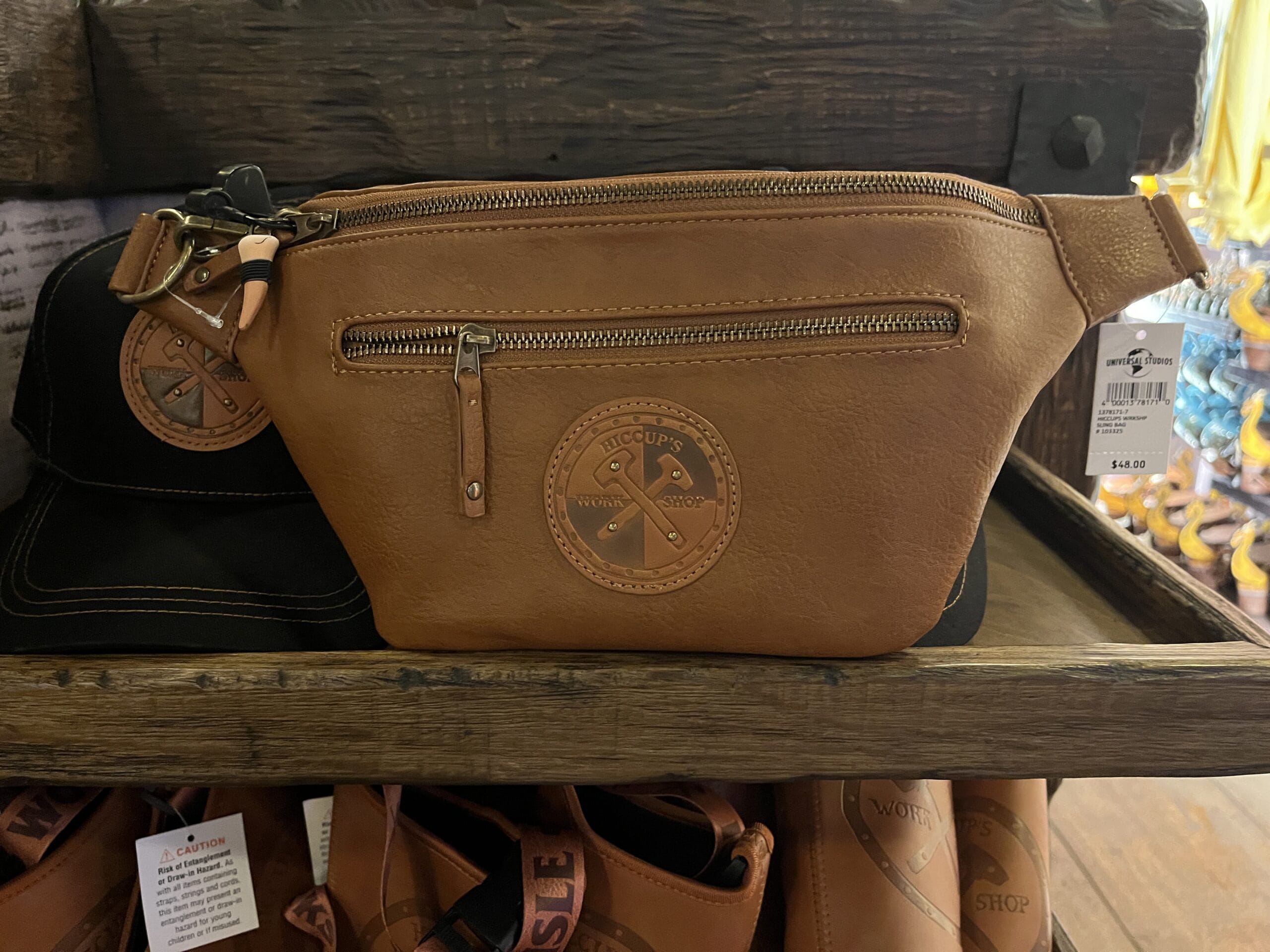 A tan leather fanny pack from Hiccup's Work Shop with a zip pocket and logo displayed on a wooden shelf.