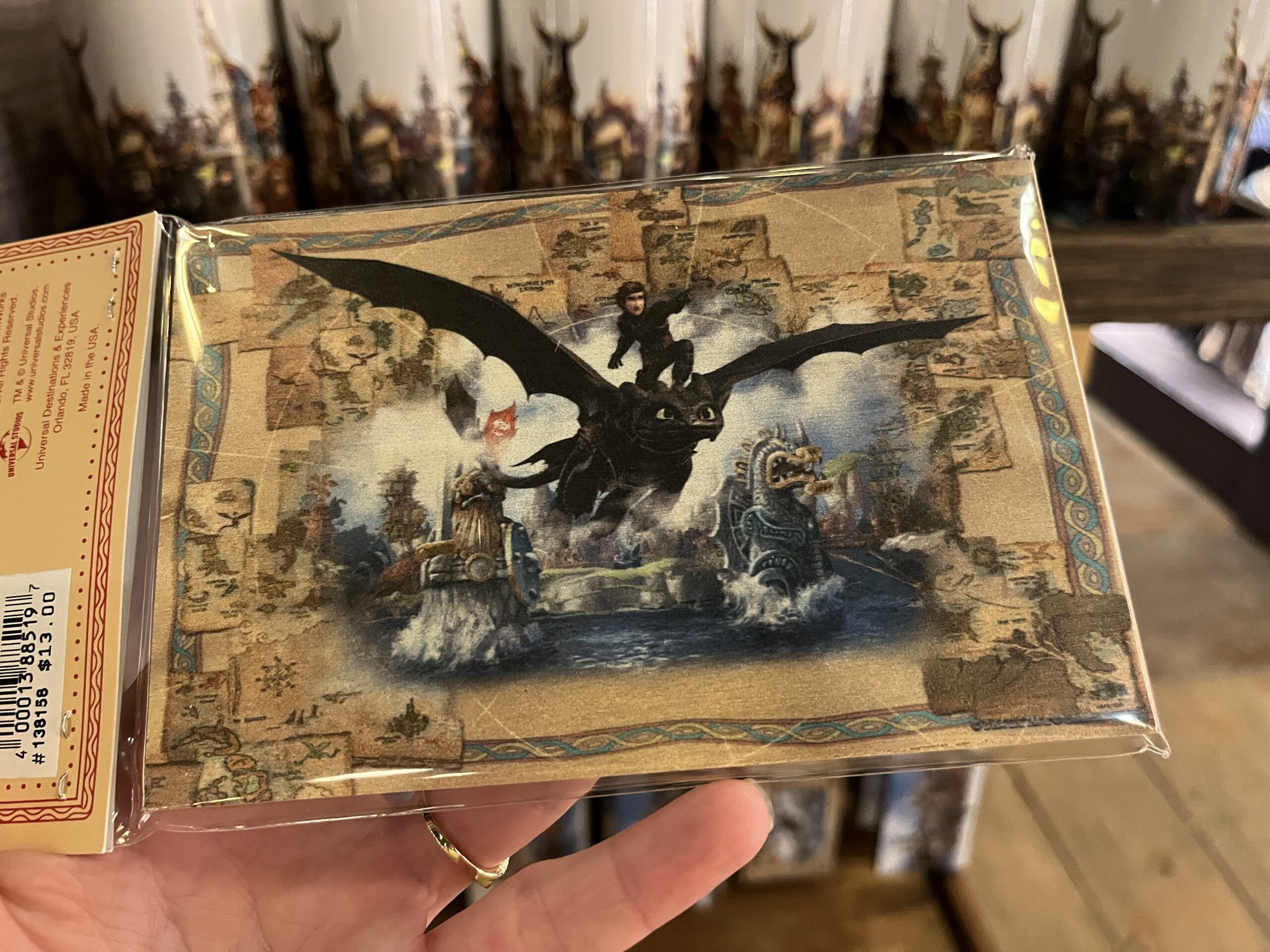 A hand holds a postcard from Hiccup's Work Shop showing a rider on a black dragon over a fantasy map with castles and water.