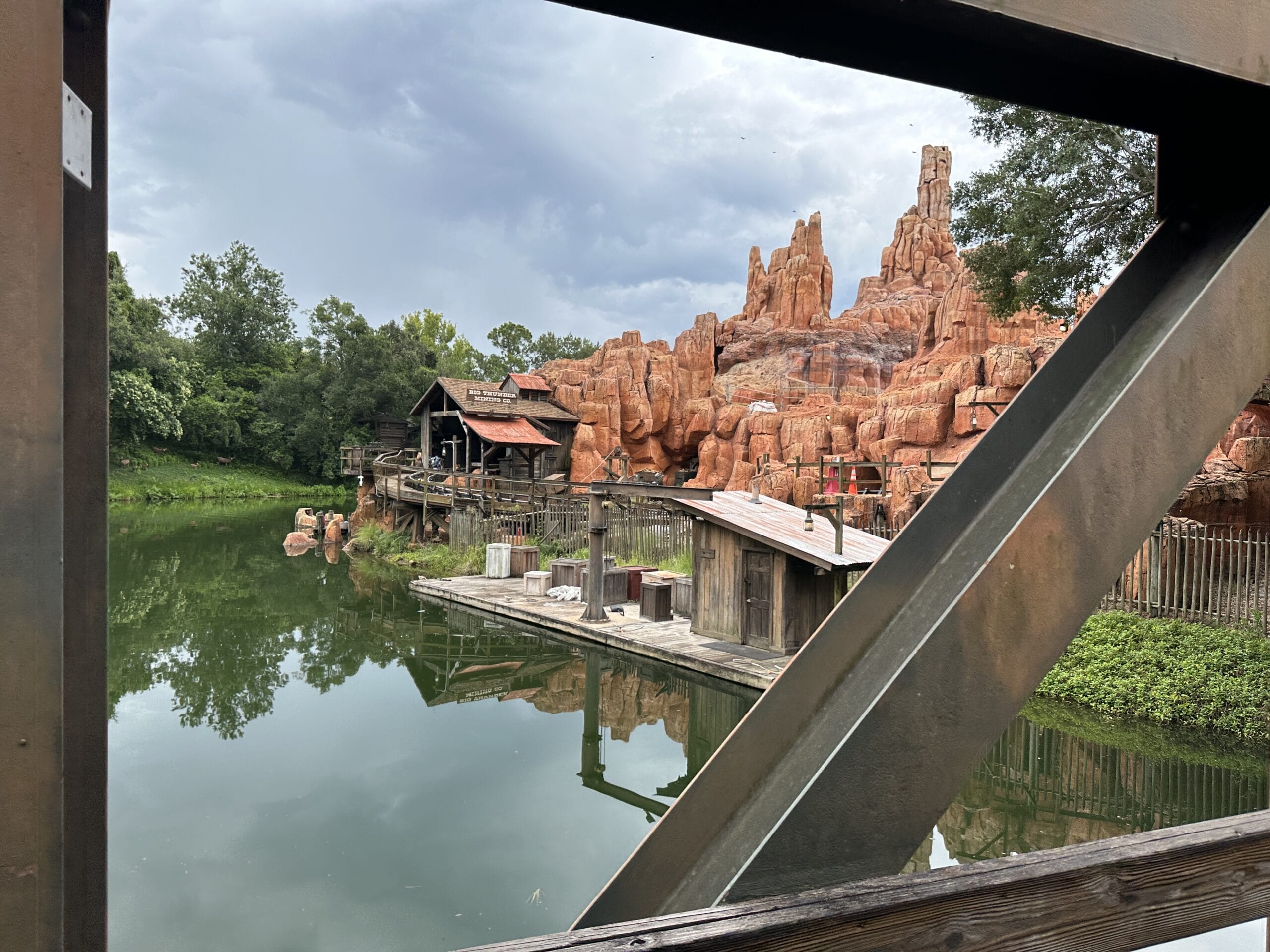 Rustic wooden buildings and rocky peaks reflect in a calm lake, near Frontierland Station on the Walt Disney World Railroad.