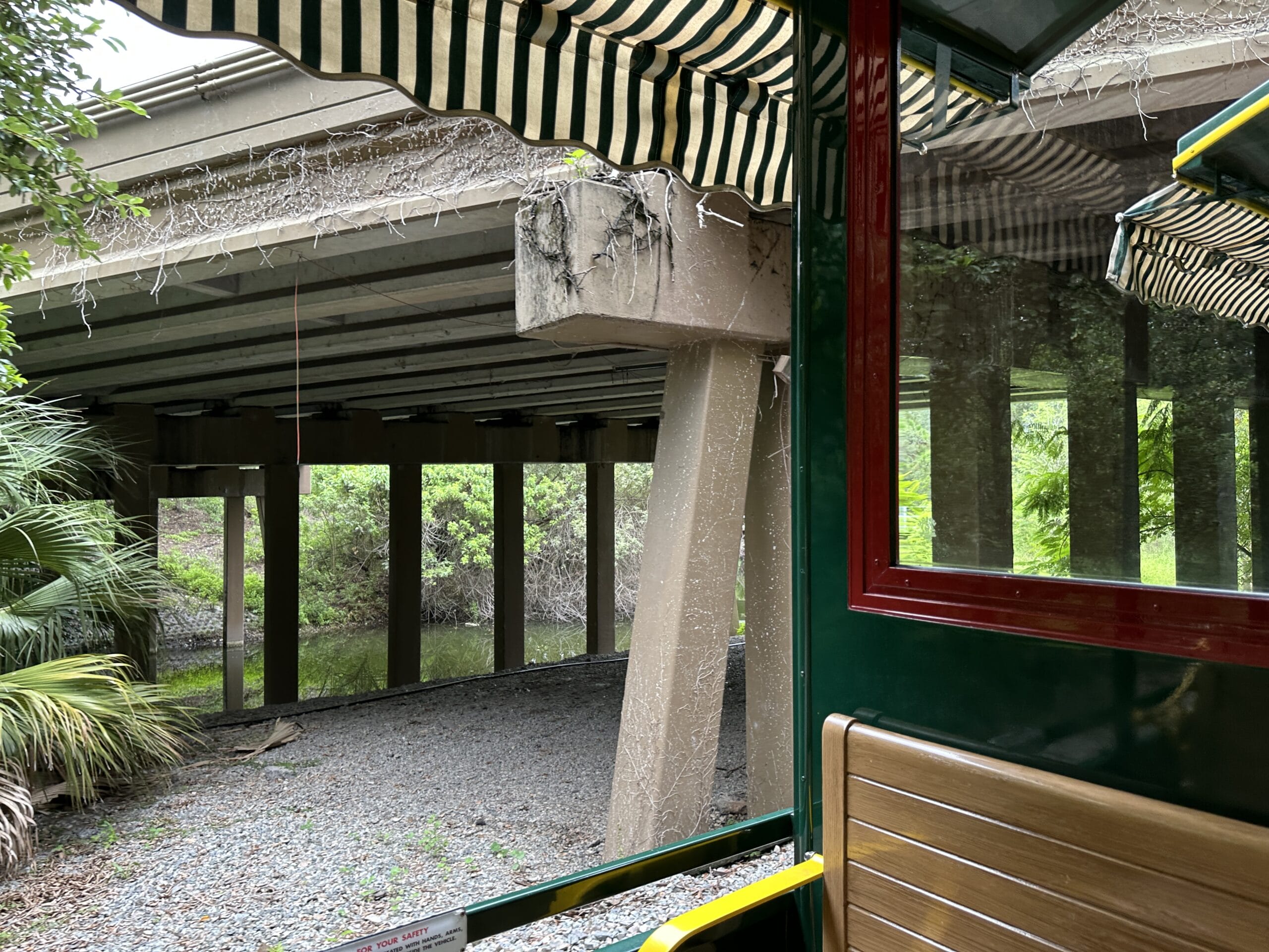 View from a train car with striped canopy on the Walt Disney World Railroad, passing under a bridge amid greenery.