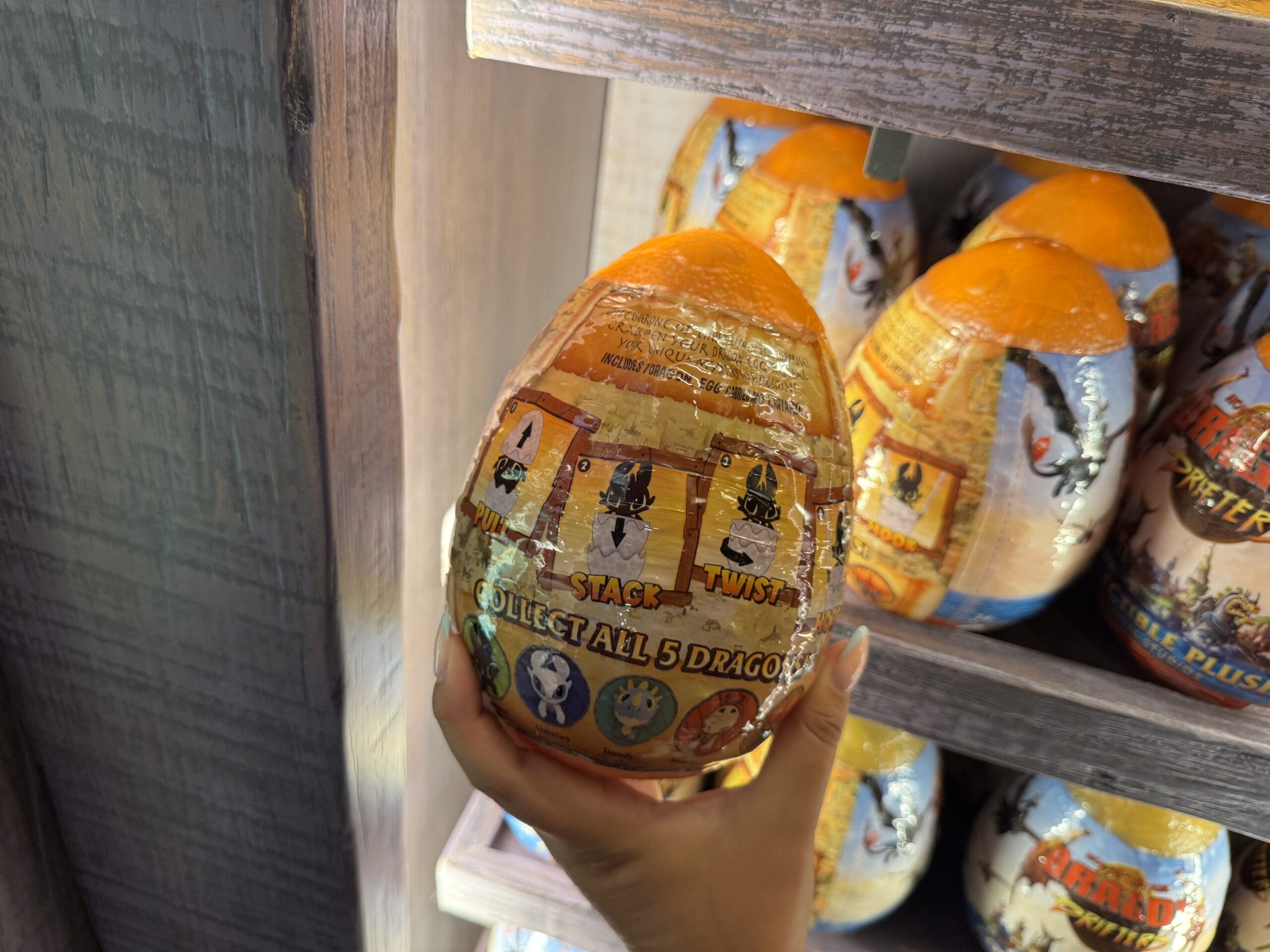 A hand holds a Hiccup's Work Shop toy egg labeled "Collect all 5 dragons" on a shelf with similar eggs.