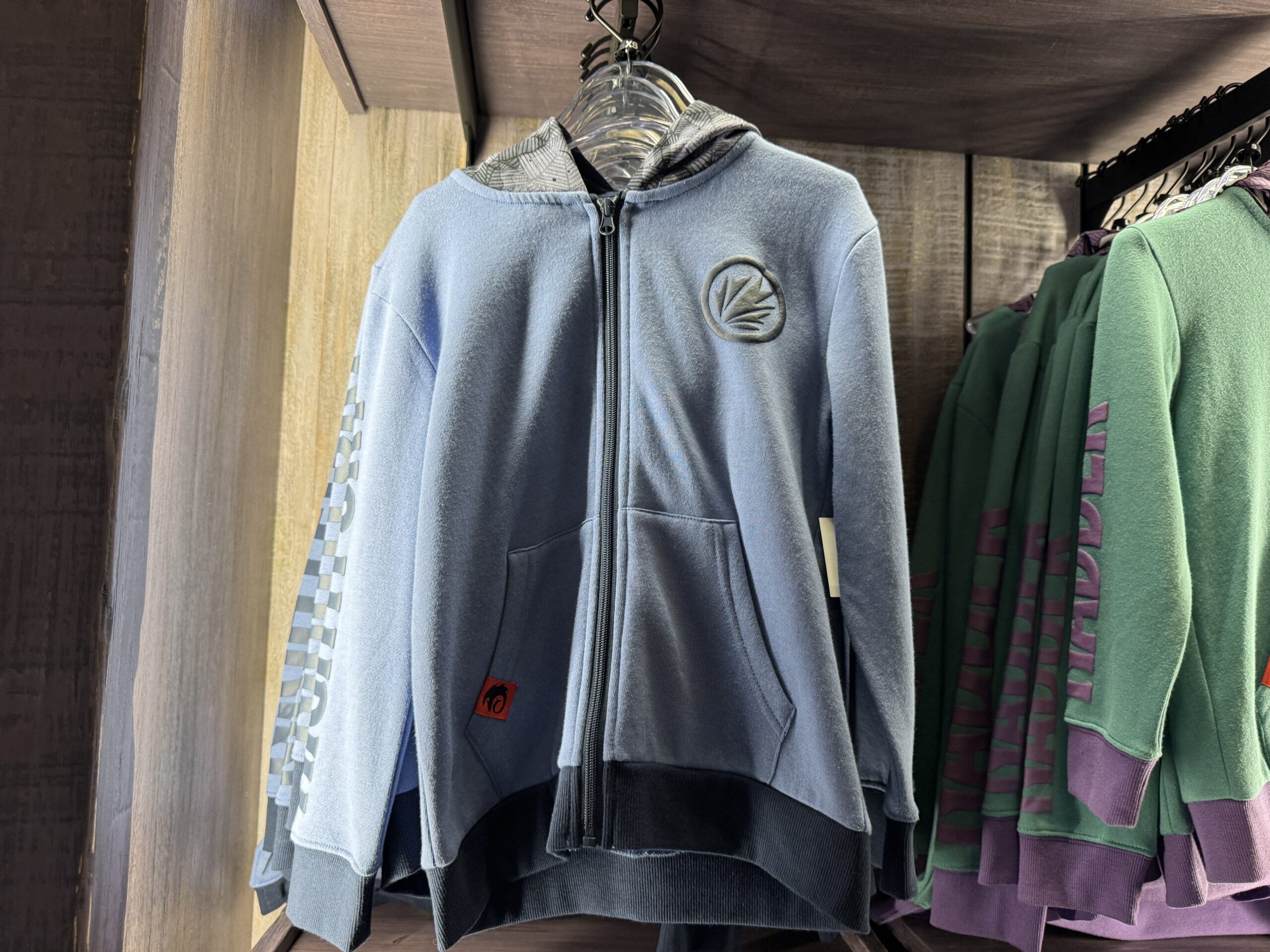 Light blue zip-up hoodie from Hiccup's Work Shop, with a round chest patch, hanging beside green hoodies in-store.