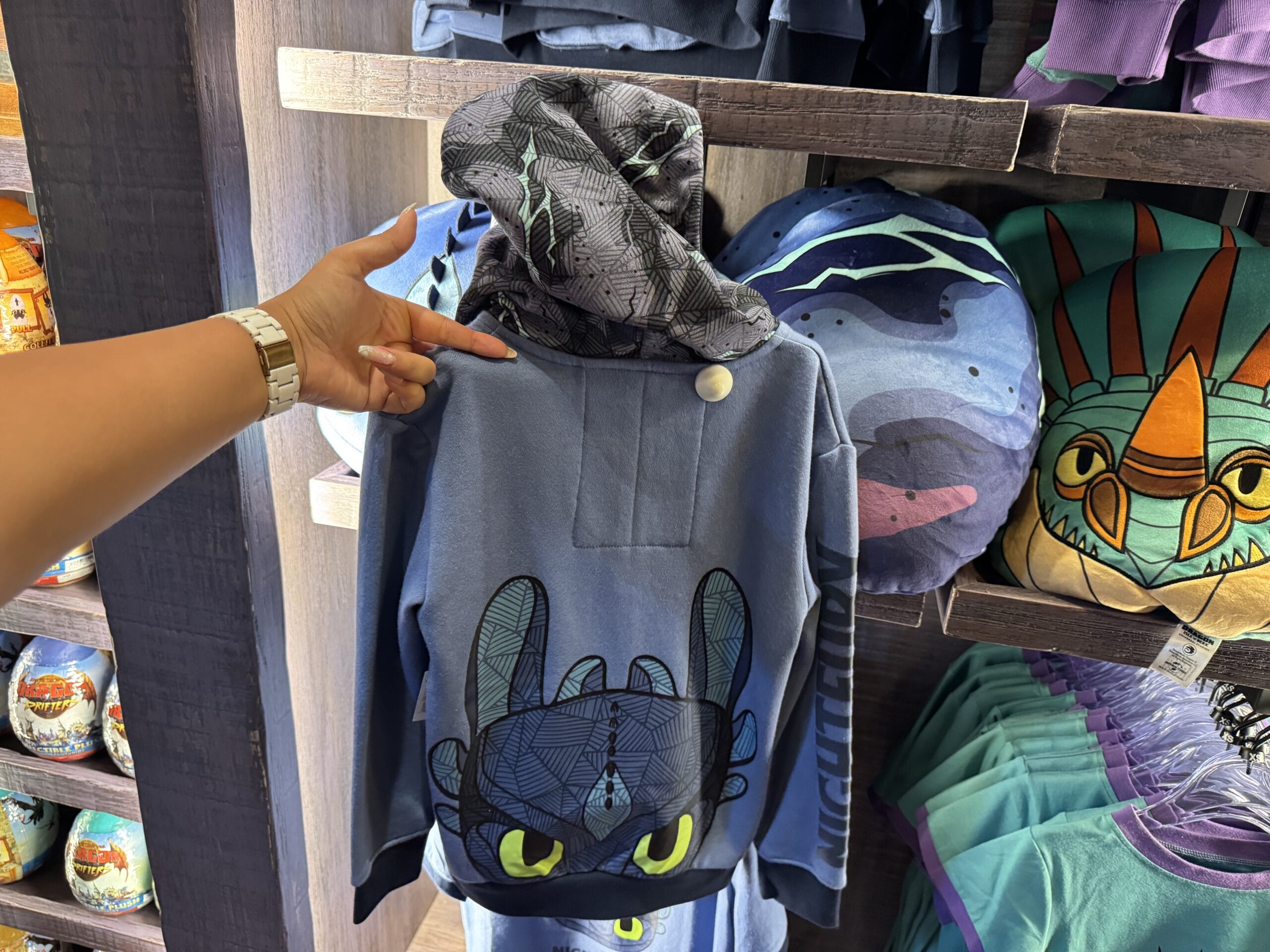 A hand points to a blue hoodie with a Toothless dragon face from Hiccup's Work Shop, displayed in a store.