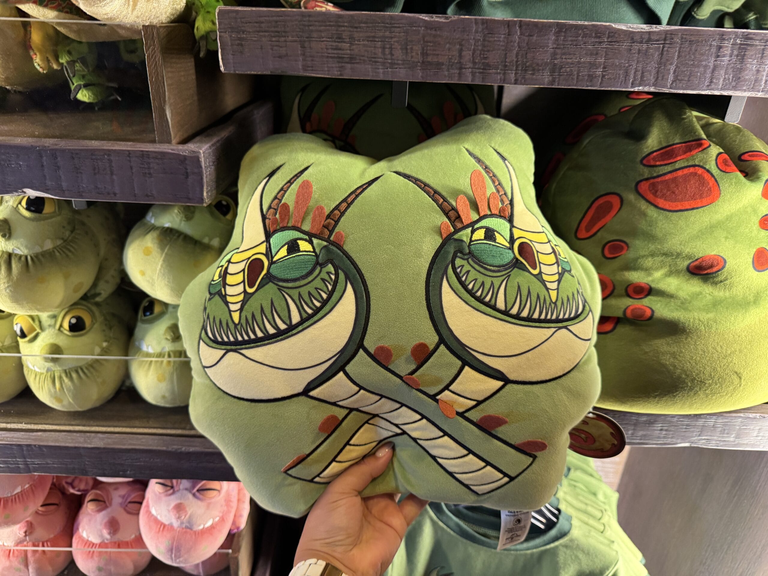 Hand holding a green pillow with two cartoonish snake heads at Hiccup's Work Shop, among plush toys on store shelves.