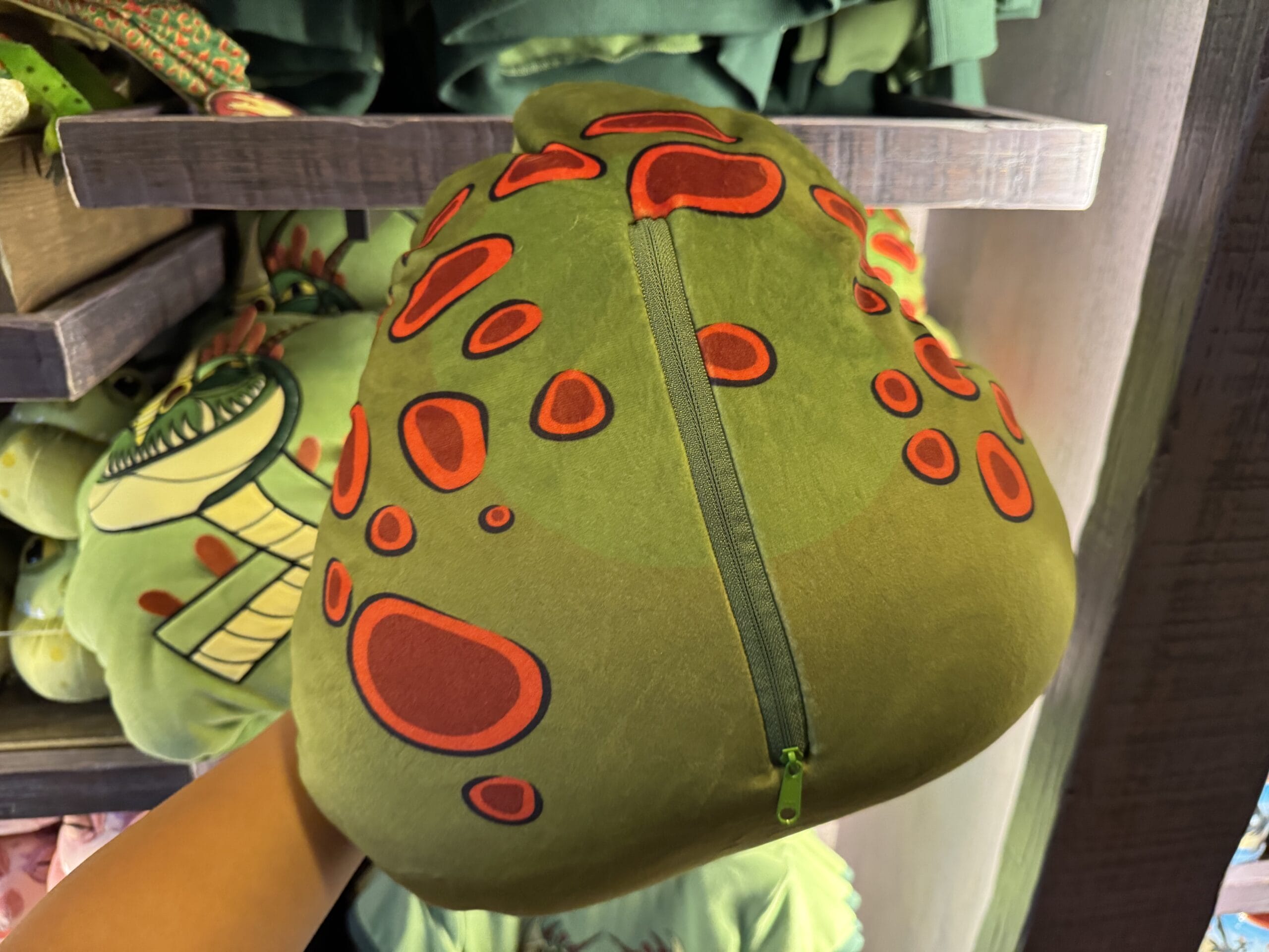 A hand holds a green, plush pillow with red spots and a zipper from Hiccup's Work Shop, displayed on a store shelf.