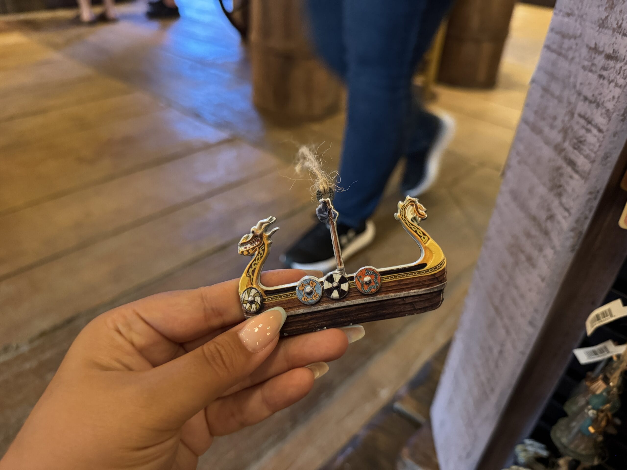 Hand holding a small Viking ship souvenir from Hiccup's Work Shop; blurred wooden floor and people in background.
