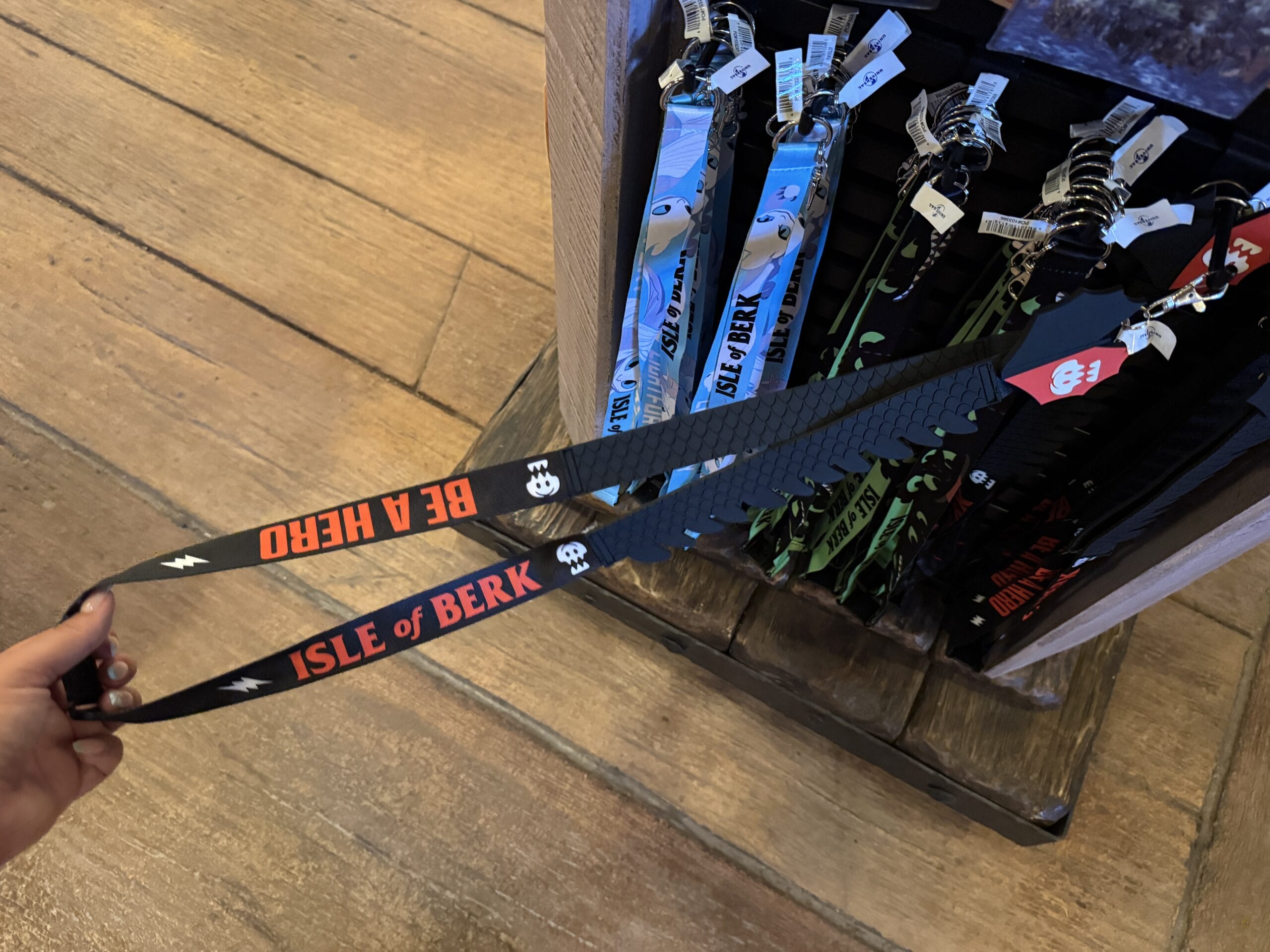 A hand holds a black "Isle of Berk" lanyard from Hiccup's Work Shop, displayed among others in a store.