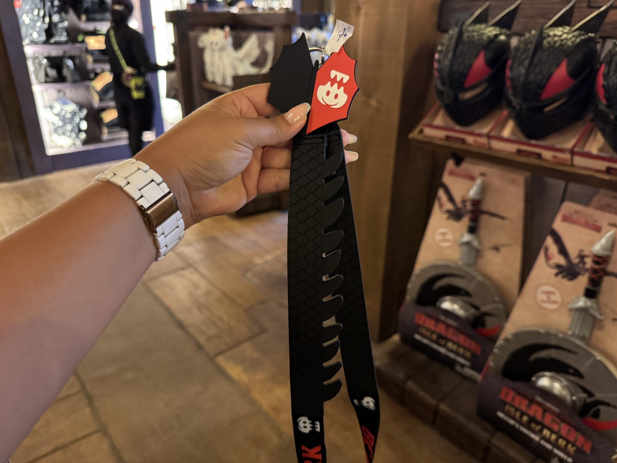A hand holds a black Mario-themed lanyard in a store near dragon helmets at Hiccup's Work Shop.