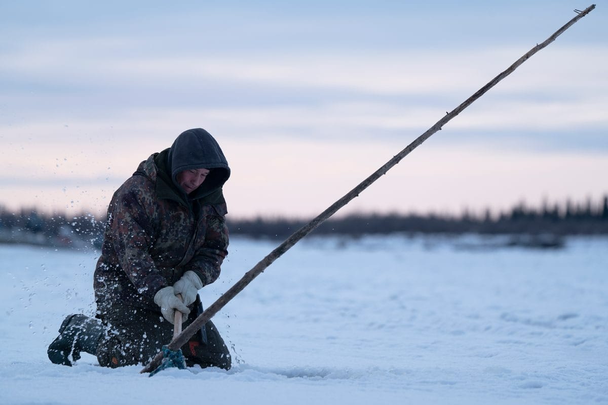 Person in winter clothing kneels on snow, using a long pole to break ice, reminiscent of an Emmy Award-worthy scene at dusk.