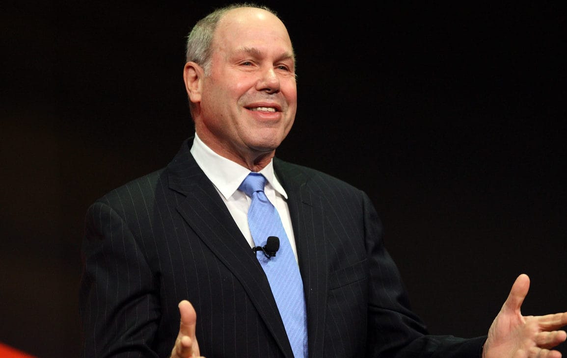 Michael Eisner in a suit and blue tie gestures while speaking on stage about Walt Disney World history.