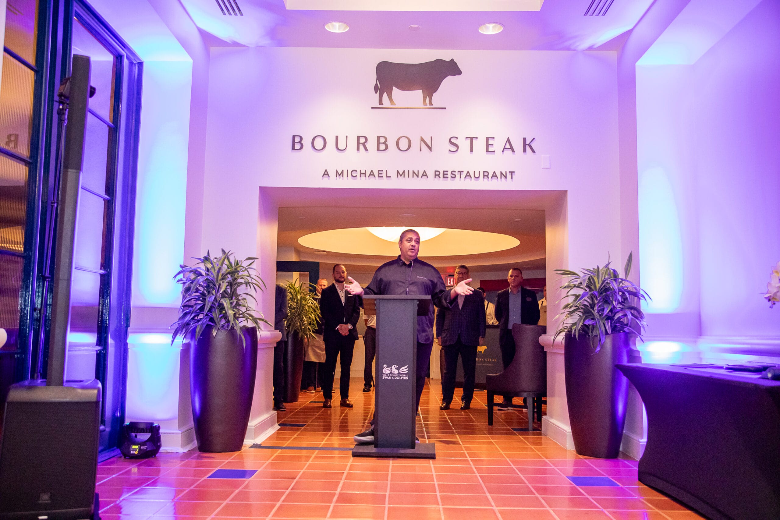 A man speaks at a podium in front of Michael Mina’s Bourbon Steak Orlando, with people behind him under purple lighting.