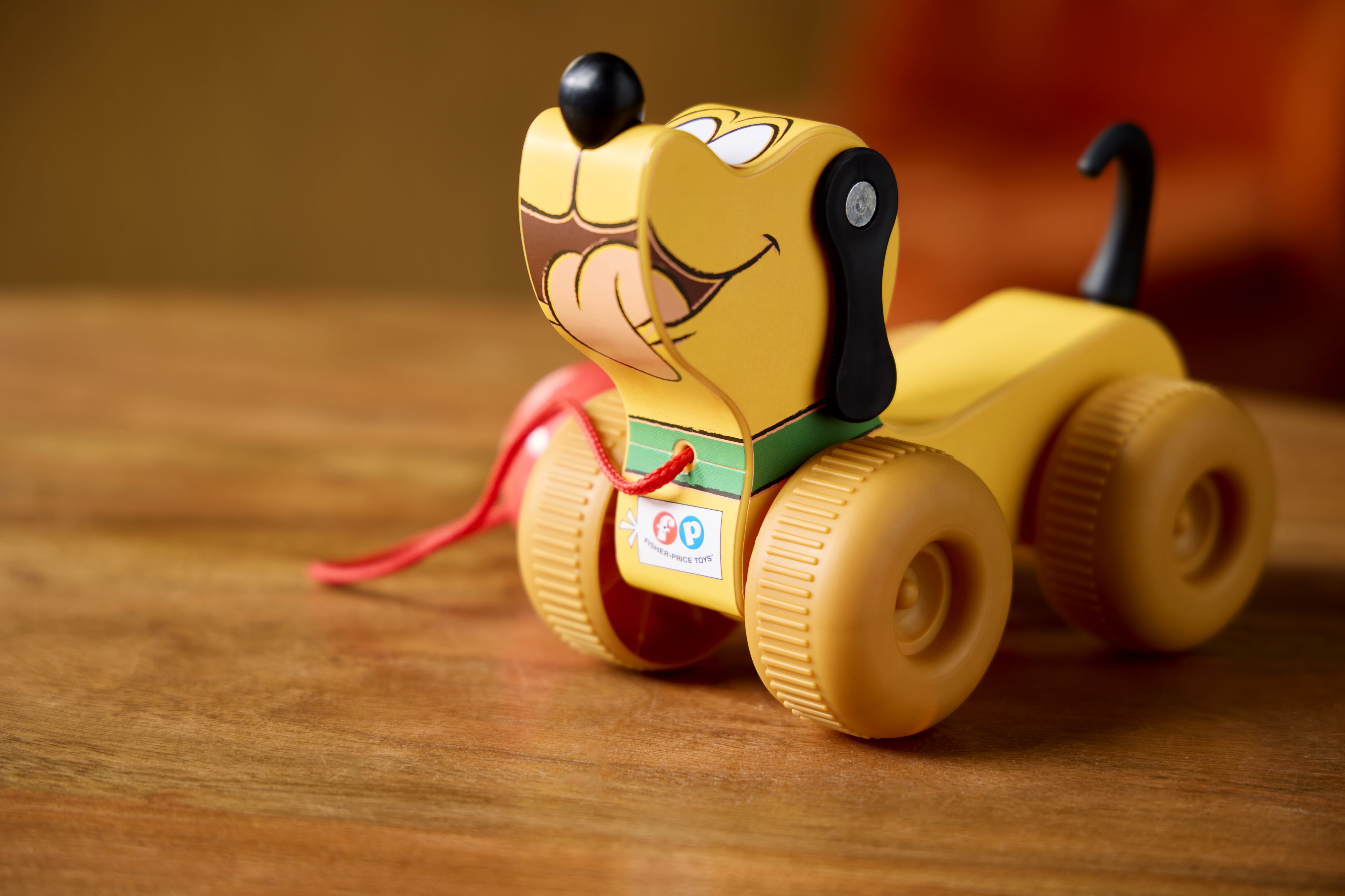 A yellow toy dog with wheels and a red pull string, inspired by Retro Fisher Price toys, sits on a wooden surface.