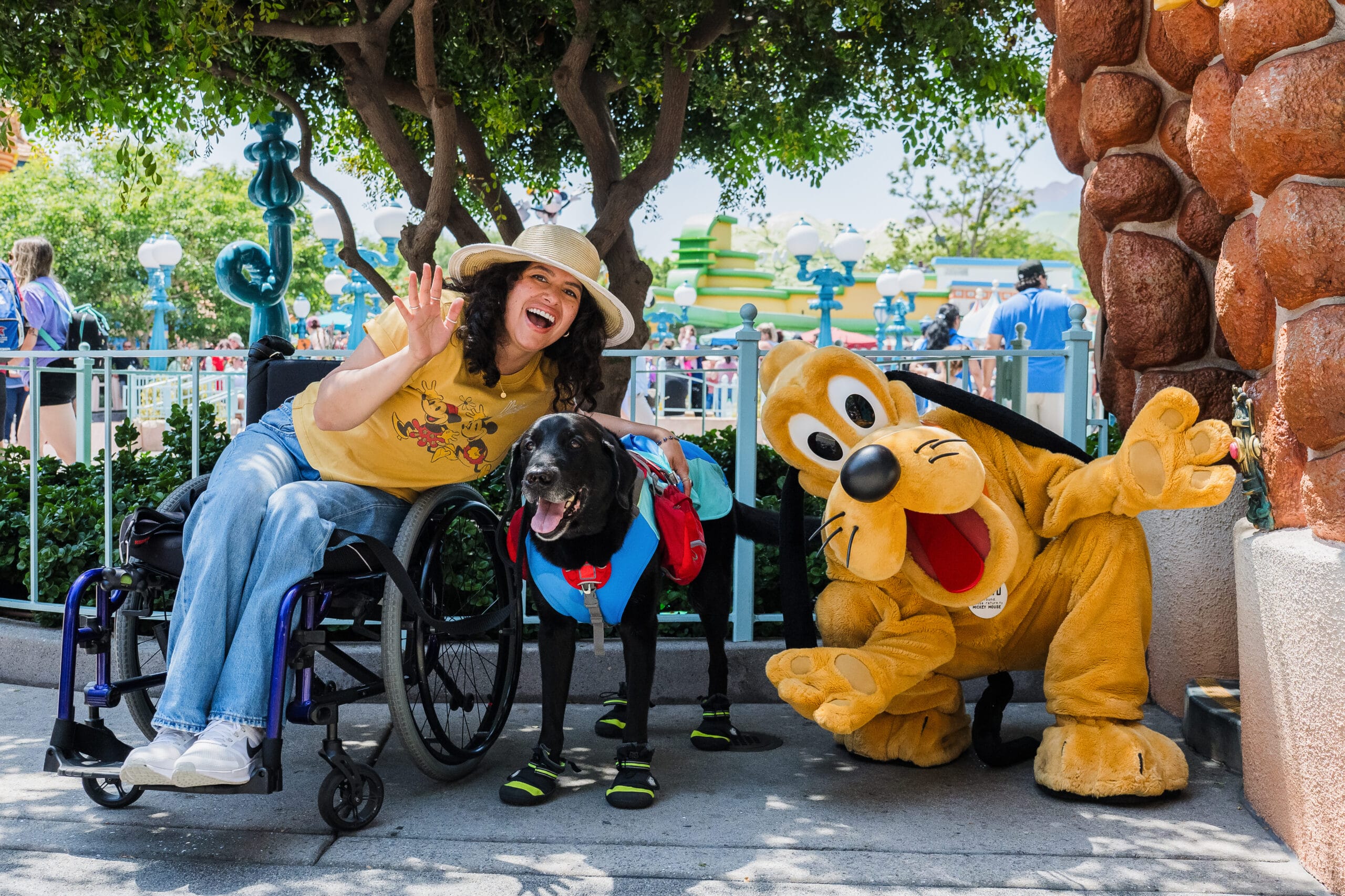 A woman in a wheelchair, her service dog, and a Pluto mascot wave and smile outside at Disneyland, the Happiest Place on Earth.