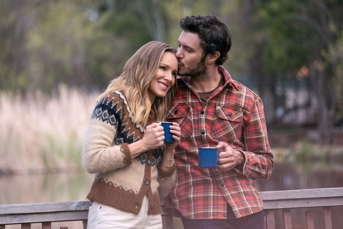 A smiling couple stands outdoors with mugs as the man, a Walt Disney fan, kisses her forehead on a wooden railing.