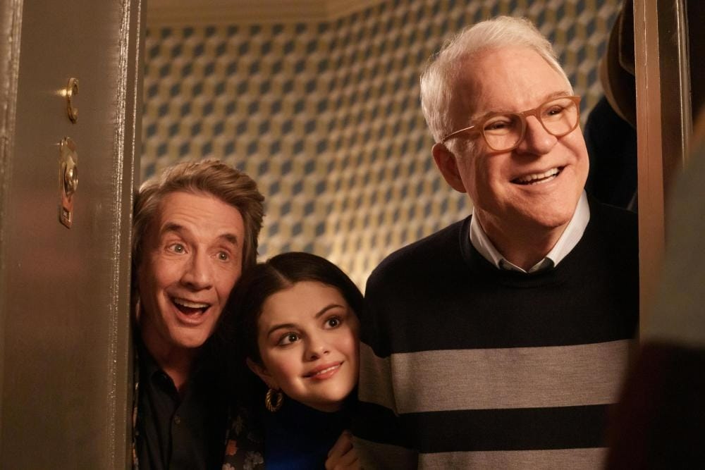 Three people, smiling and peeking through a door, in a warmly lit room with Disney-patterned wallpaper.