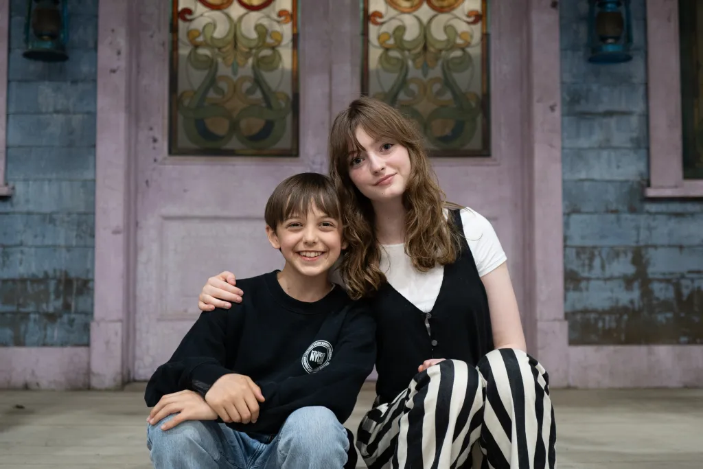 Two smiling kids sit on wooden steps, like Children of Hades from Percy Jackson Season Three, by a decorative purple door.