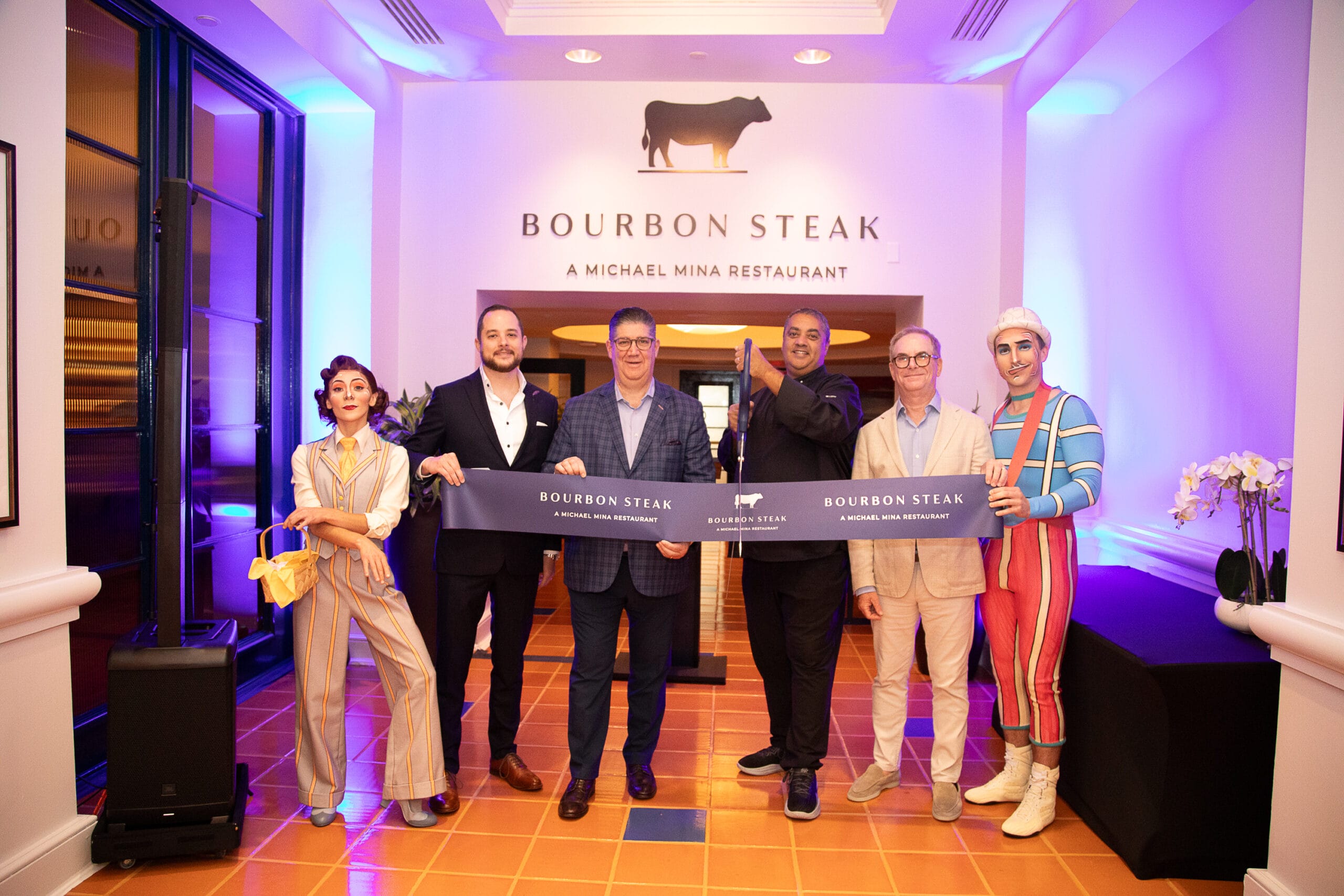 Six people stand at the Bourbon Steak Orlando entrance at Walt Disney World Dolphin Hotel, holding a ribbon for the grand opening.