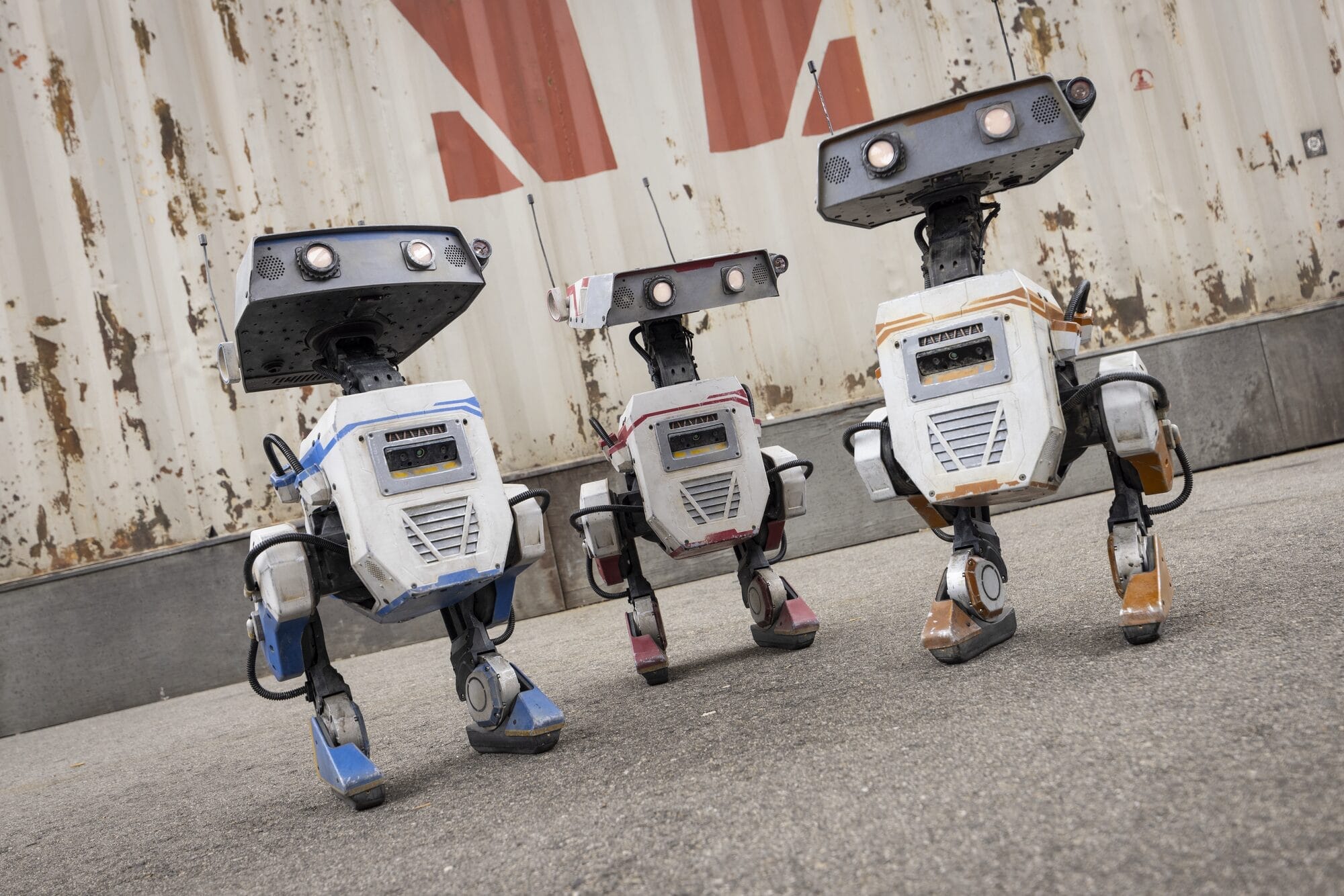 Three boxy, two-legged bdx droids with large eyes stand on pavement before a weathered shipping container.