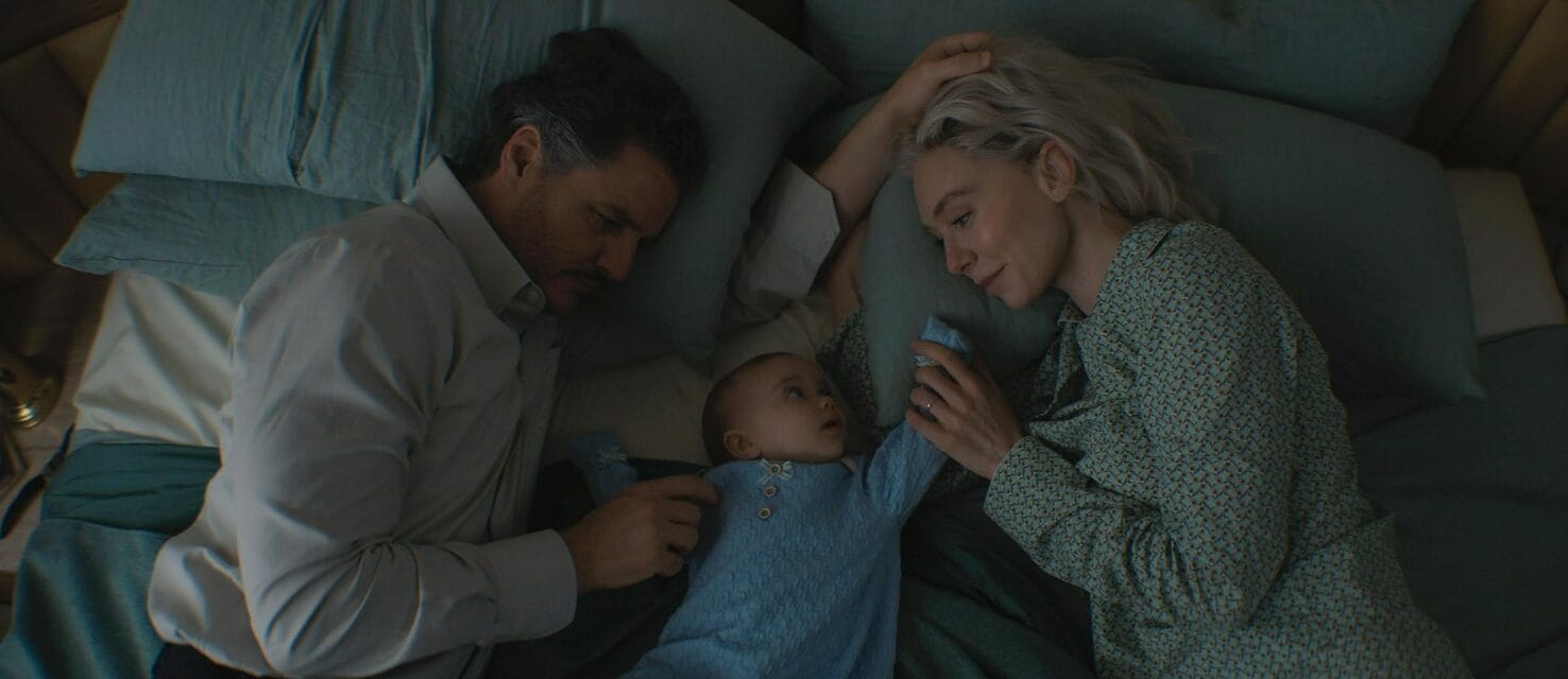 A man, woman, and baby lie together on a bed, sharing First Steps and affectionate looks in soft lighting.