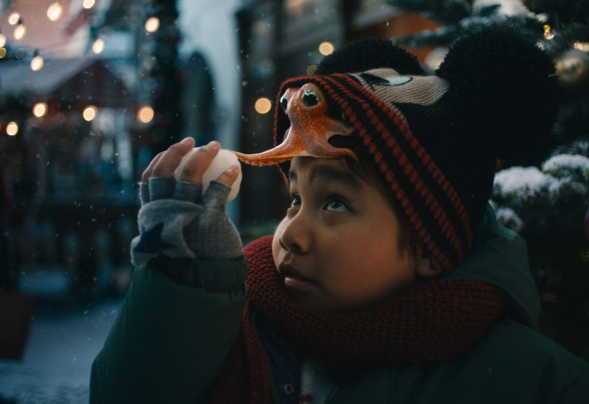 A child in winter clothes, holding a snowball, wears a hat topped with a toy fish—true Walt Disney Company charm.