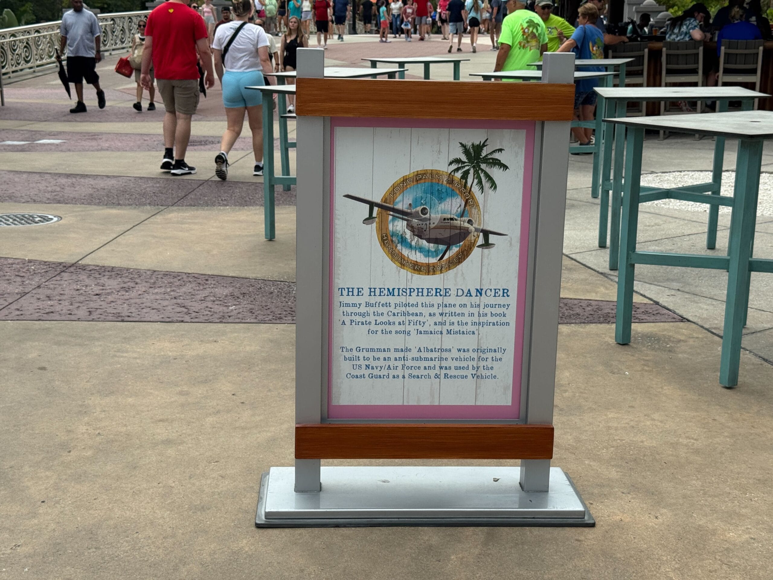 A sign about Jimmy Buffett’s Hemisphere Dancer stands on a pathway with people strolling in the background.