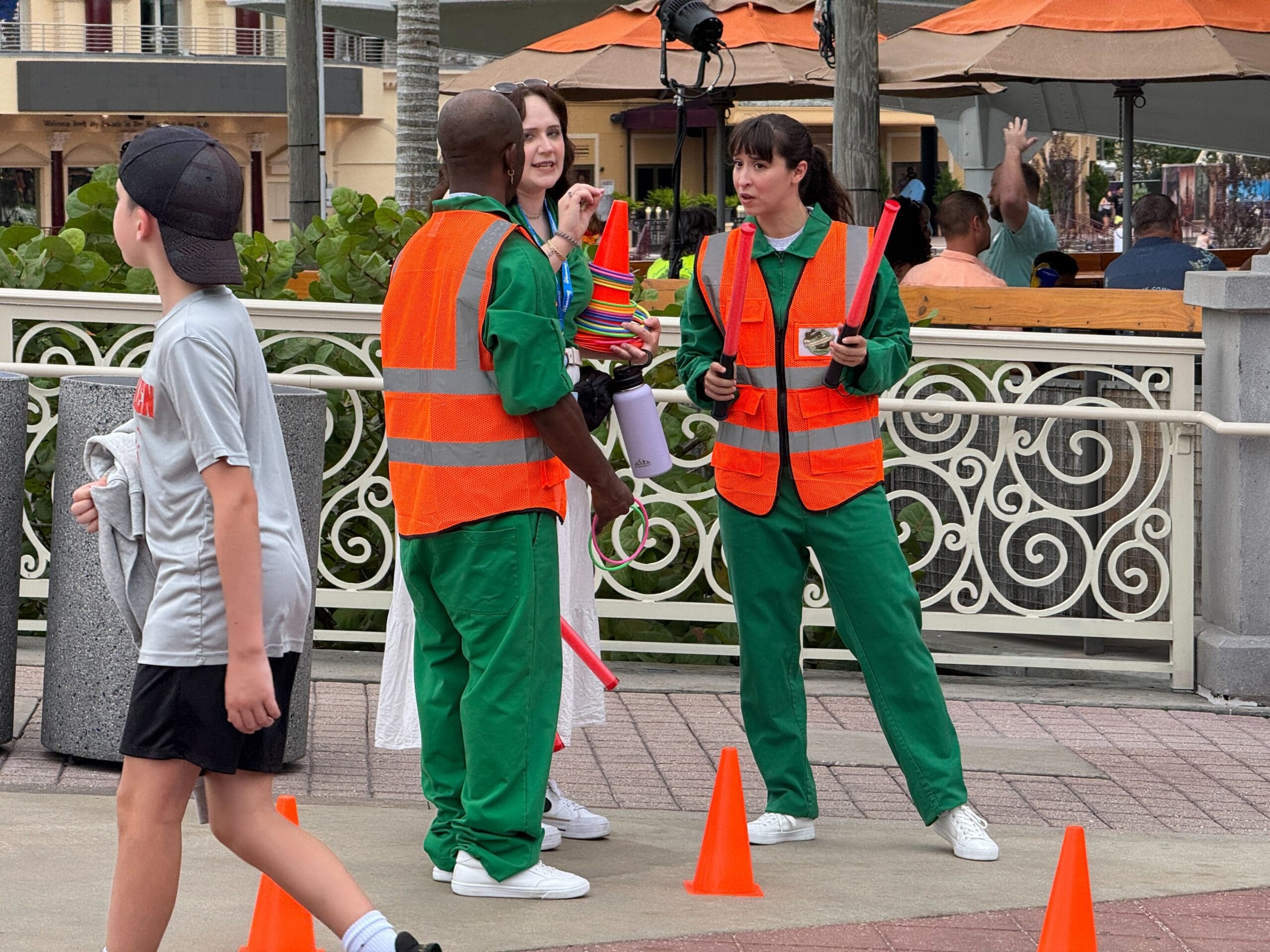 Three people in orange safety vests talk near cones as a boy walks by, recalling a Margaritaville sidewalk scene.