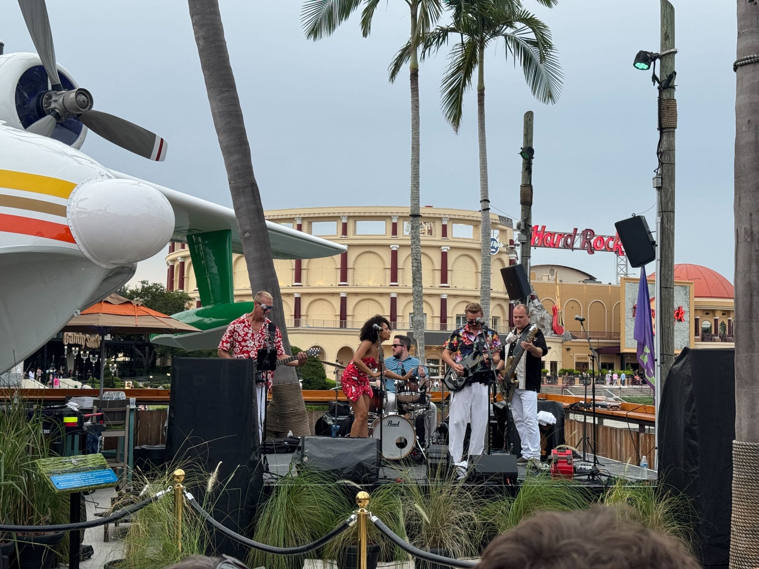 A band performs near palm trees and the Hemisphere Dancer seaplane, with a Hard Rock Cafe in the background.