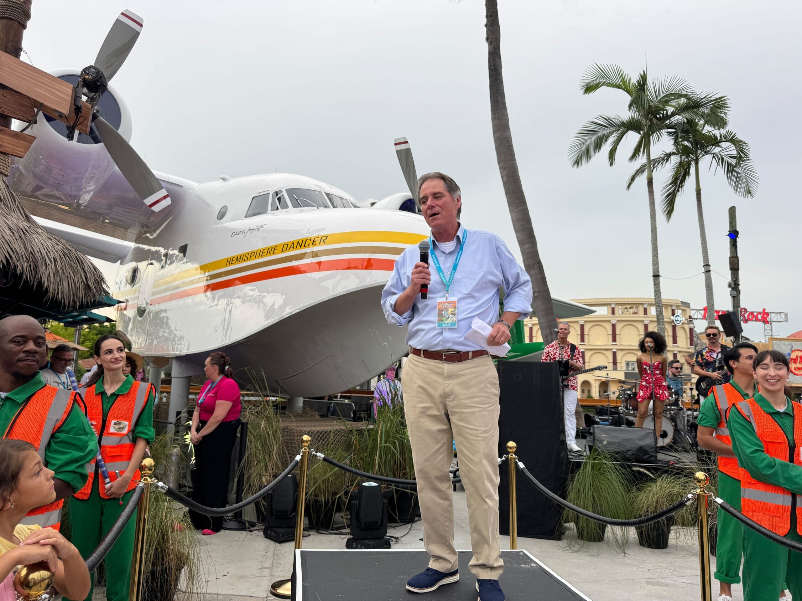 A man speaks into a microphone outdoors onstage, with Jimmy Buffett’s Hemisphere Dancer and people in orange vests nearby.