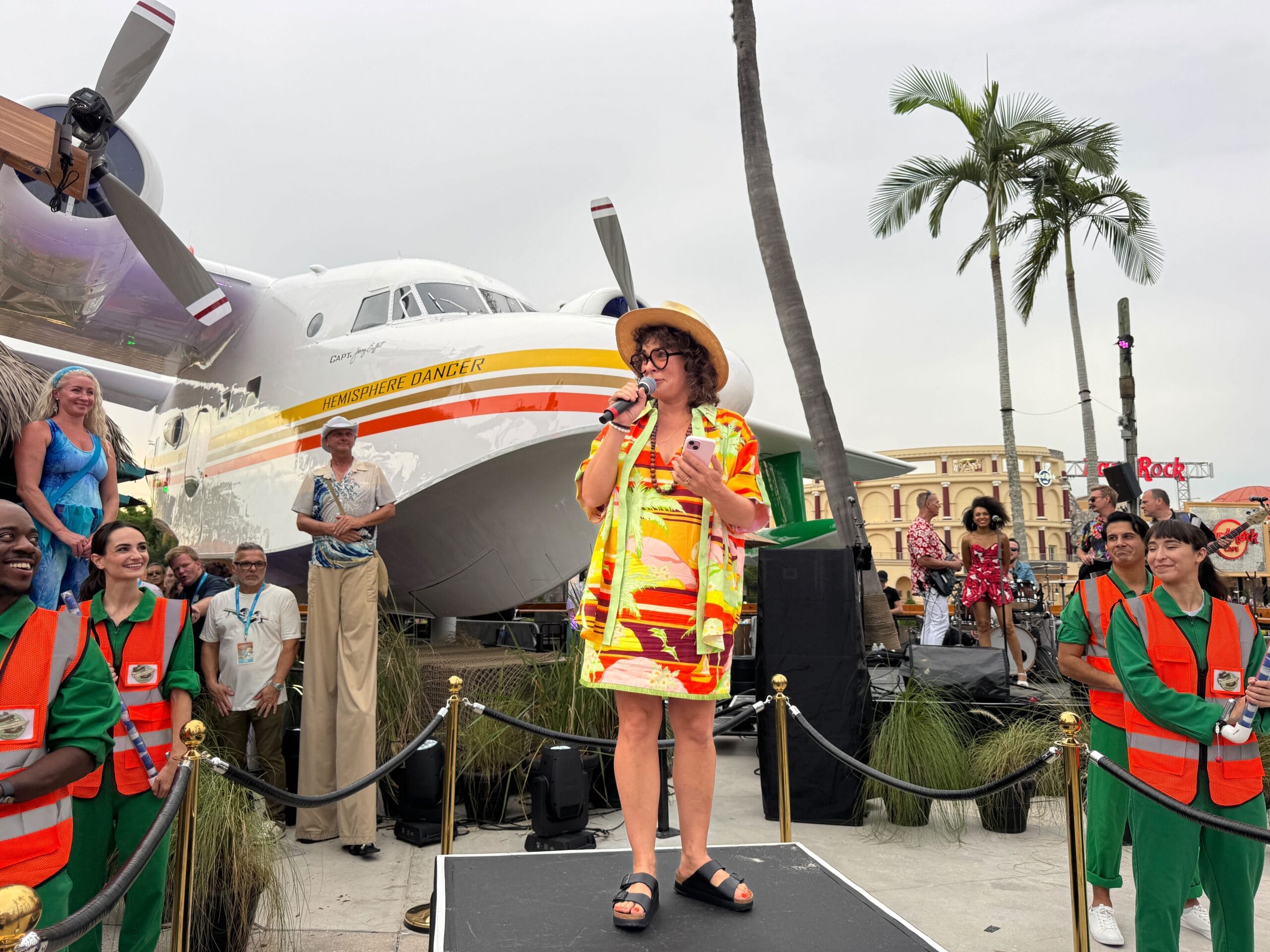 A woman in a tropical outfit speaks onstage at Universal CityWalk, framed by a seaplane and palm tree in Margaritaville style.