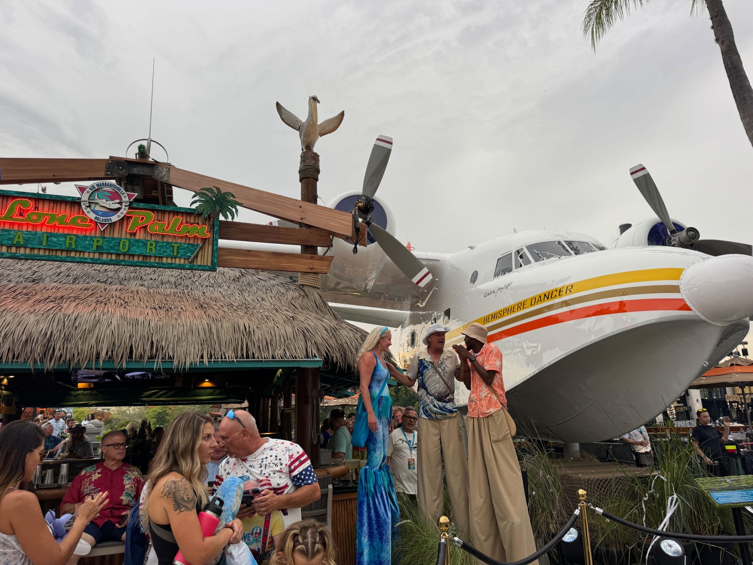 People gather at an outdoor tiki bar near Jimmy Buffett’s Hemisphere Dancer, with performers on stilts and palm trees around.