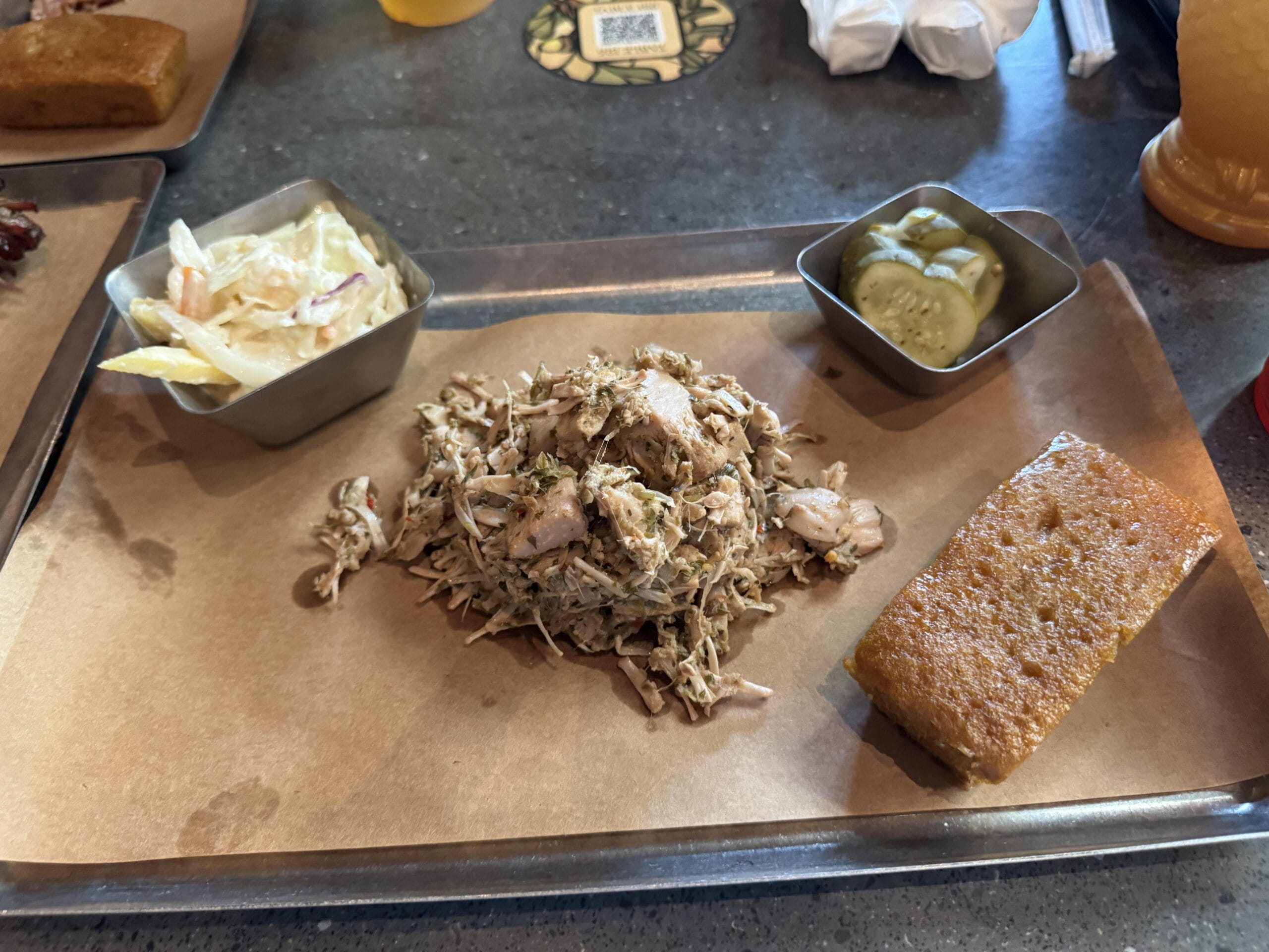 A tray from The Oak & Star Tavern with pulled pork, cornbread, coleslaw, and pickles on brown paper.