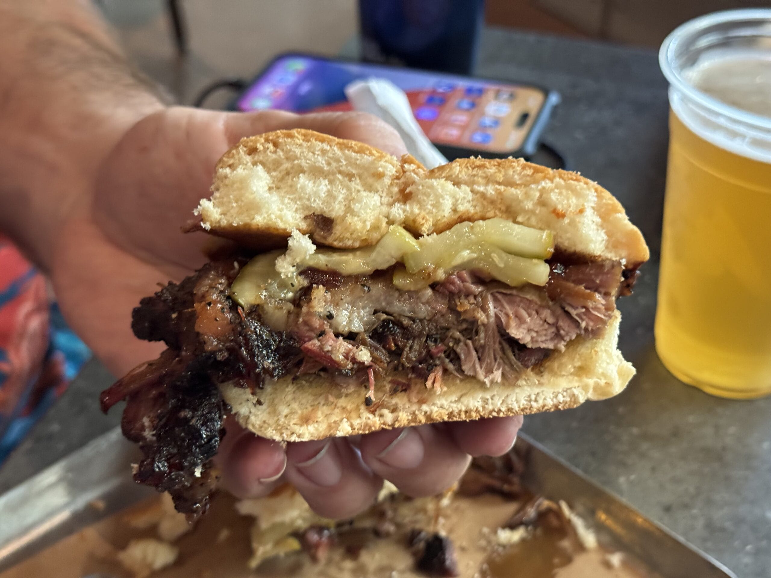 A hand holds a brisket sandwich with cheese at The Oak & Star Tavern; tray, drink, and phone in the background.