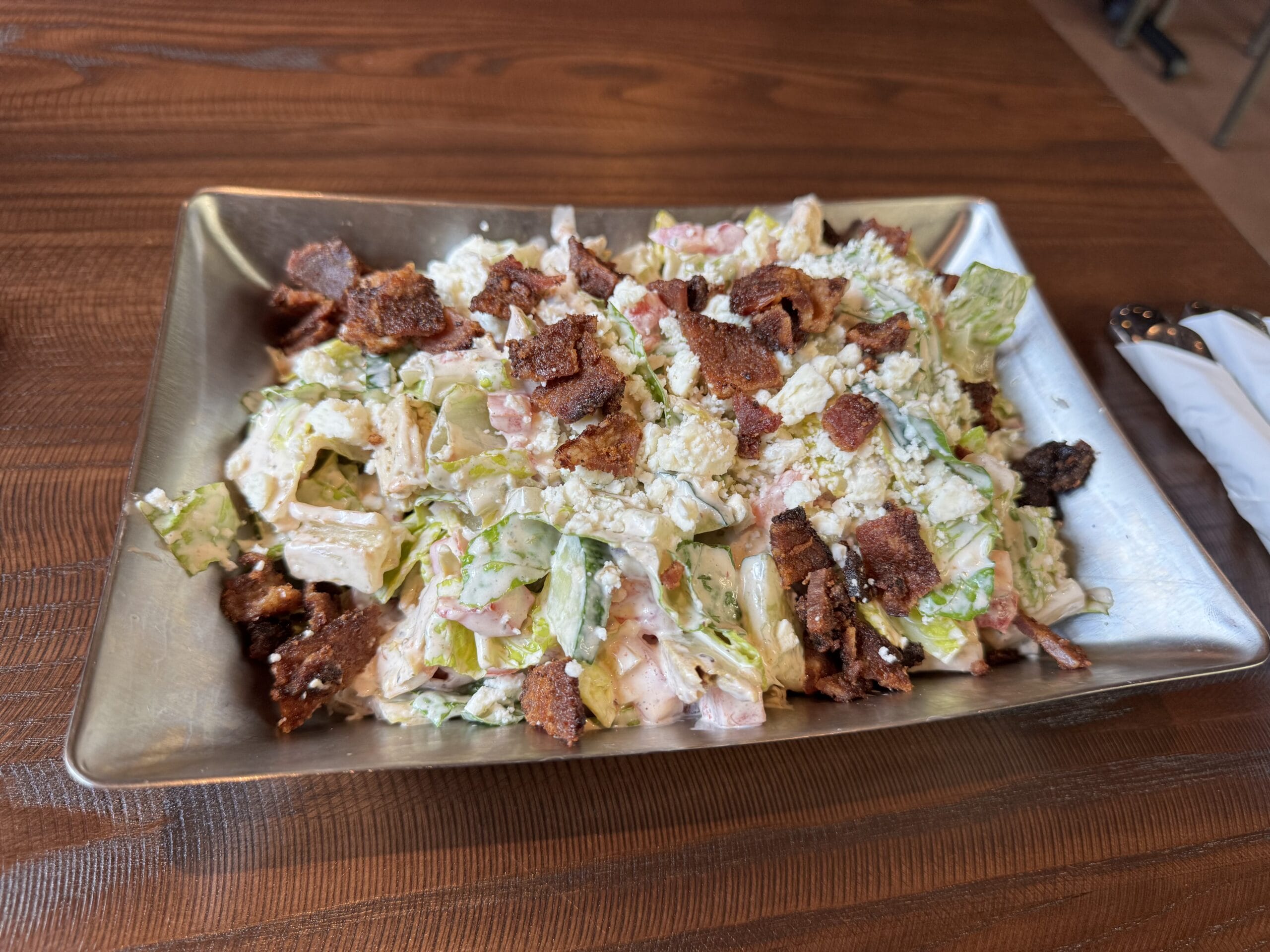 A metal tray of chopped salad from The Oak & Star Tavern, topped with crumbled cheese and crispy bacon pieces.