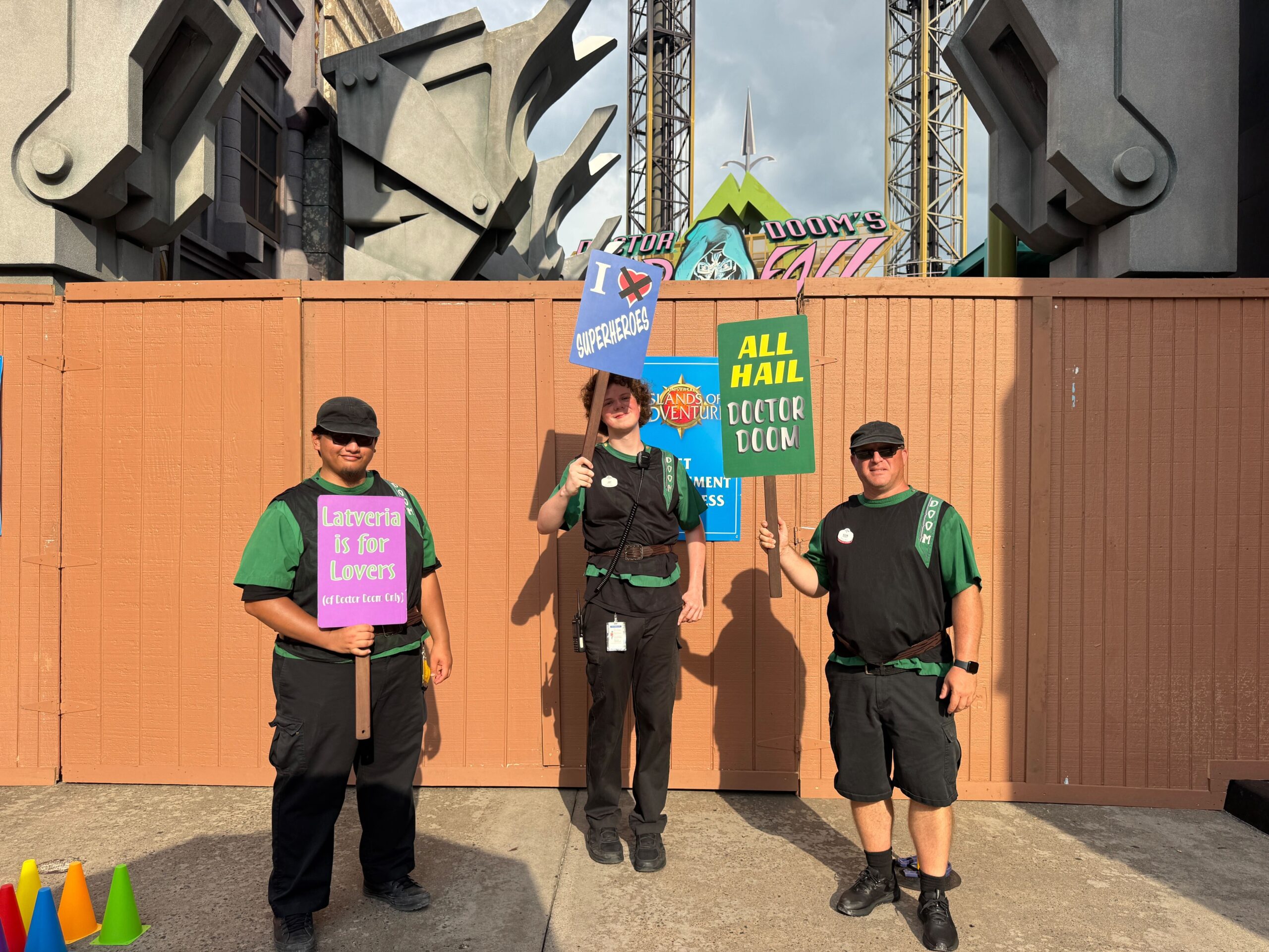 Three people in black and green uniforms hold humorous protest signs, referencing Reed Richards, at a closed amusement park attraction.