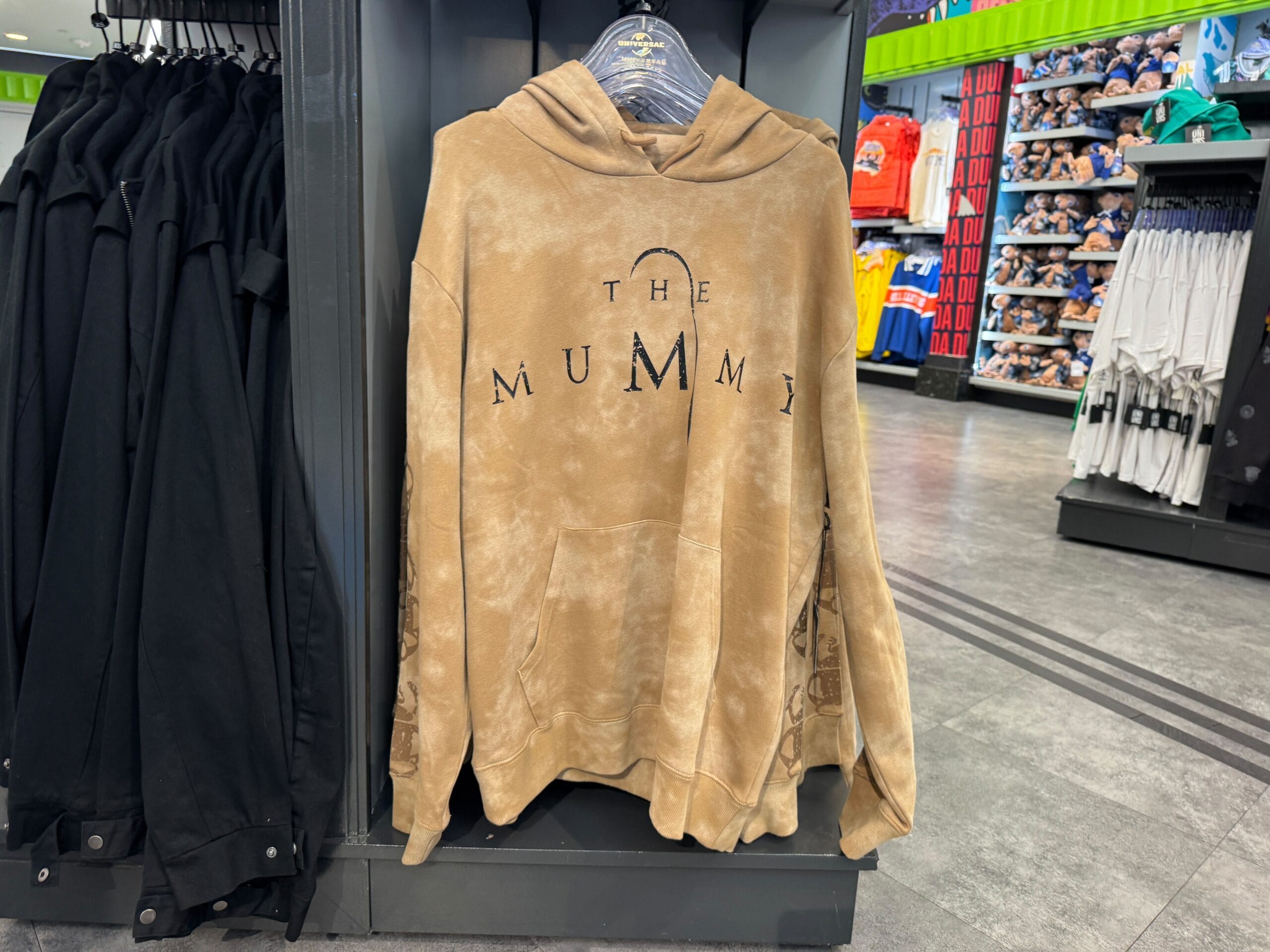 Tan "The Mummy" hoodie from the UNIVRS Mummy Collection, with black text, displayed among other apparel in-store.