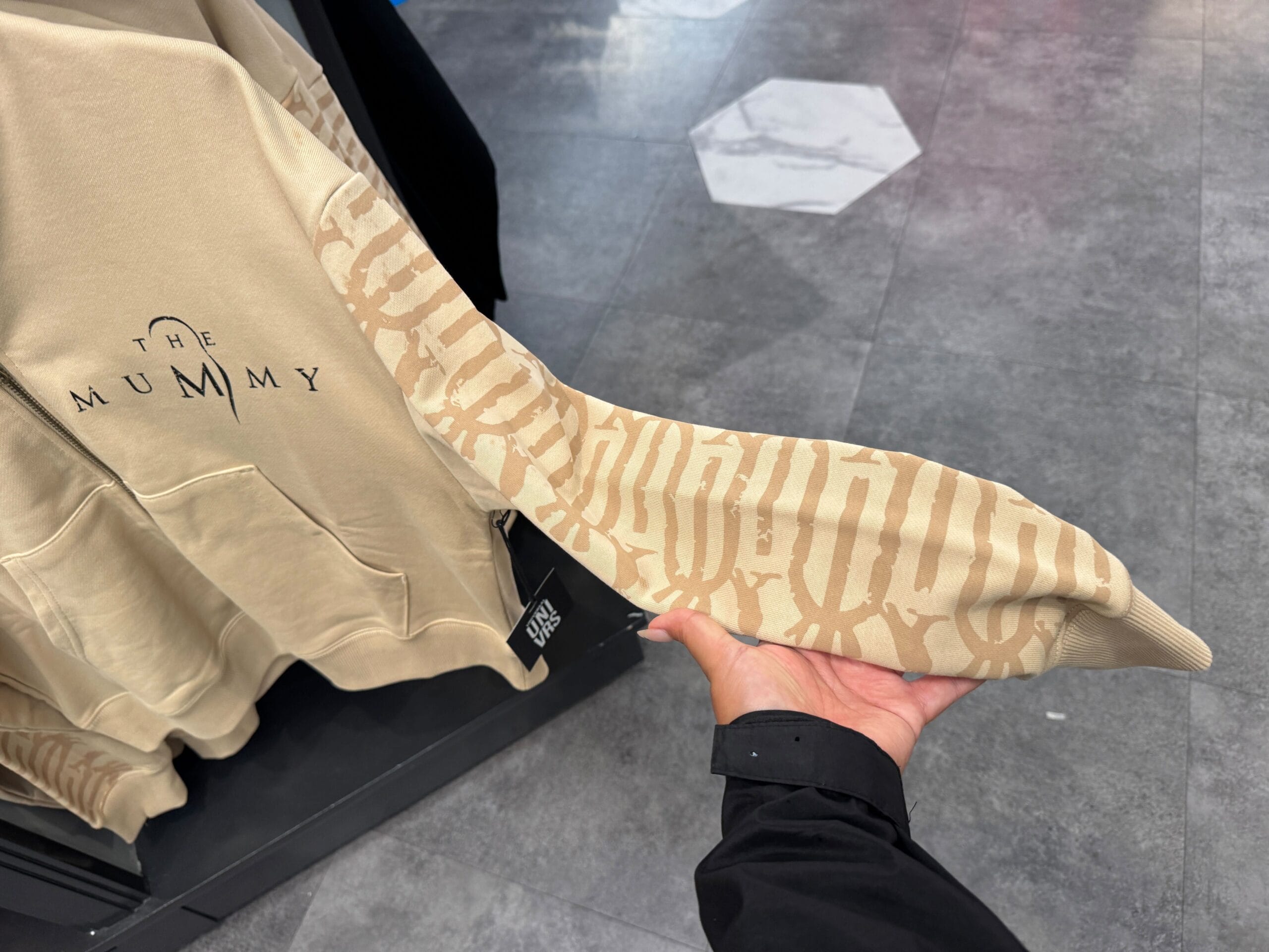 A person holds a beige hoodie sleeve from the UNIVRS Mummy Collection, featuring "The Mummy" on the chest.