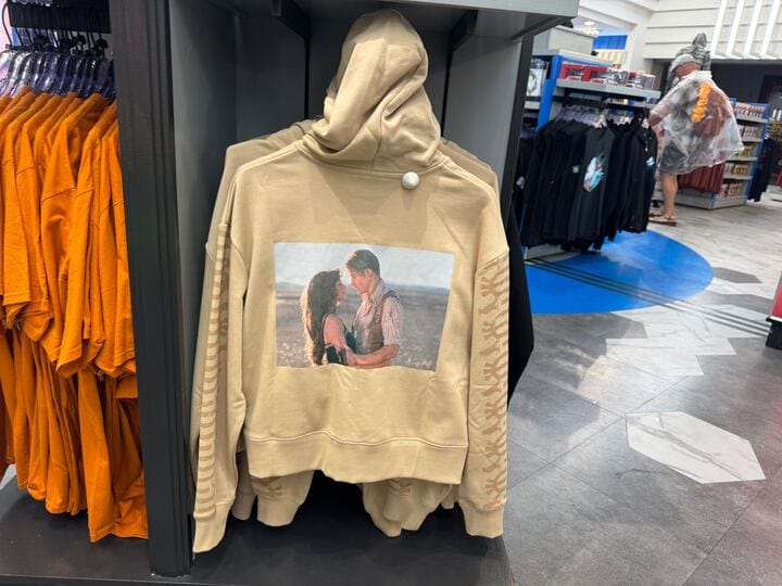 Beige hoodie from the UNIVRS Mummy Collection, featuring a couple embracing, displayed beside orange and black shirts.
